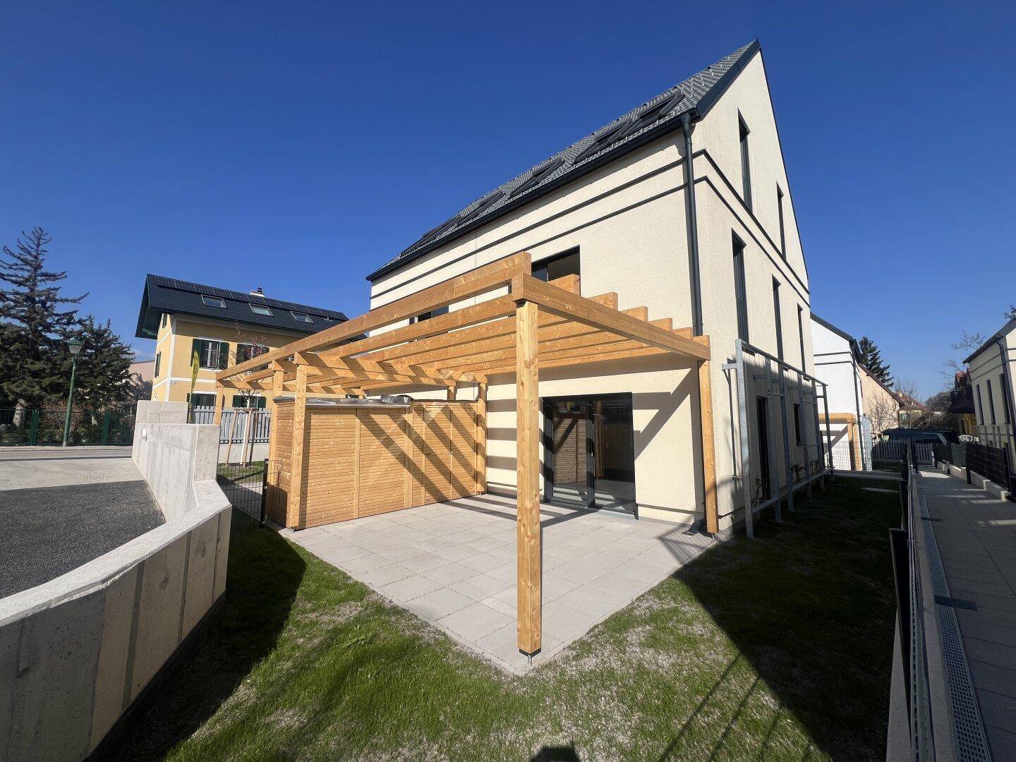 Moderne Hausfassade mit großer gepflasterter Terrasse, Holzpergola und umliegendem Rasen.