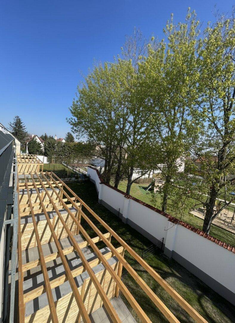 Blick von einem im Bau befindlichen Balkon auf den grünen Gartenbereich mit Bäumen und Spielplatz.