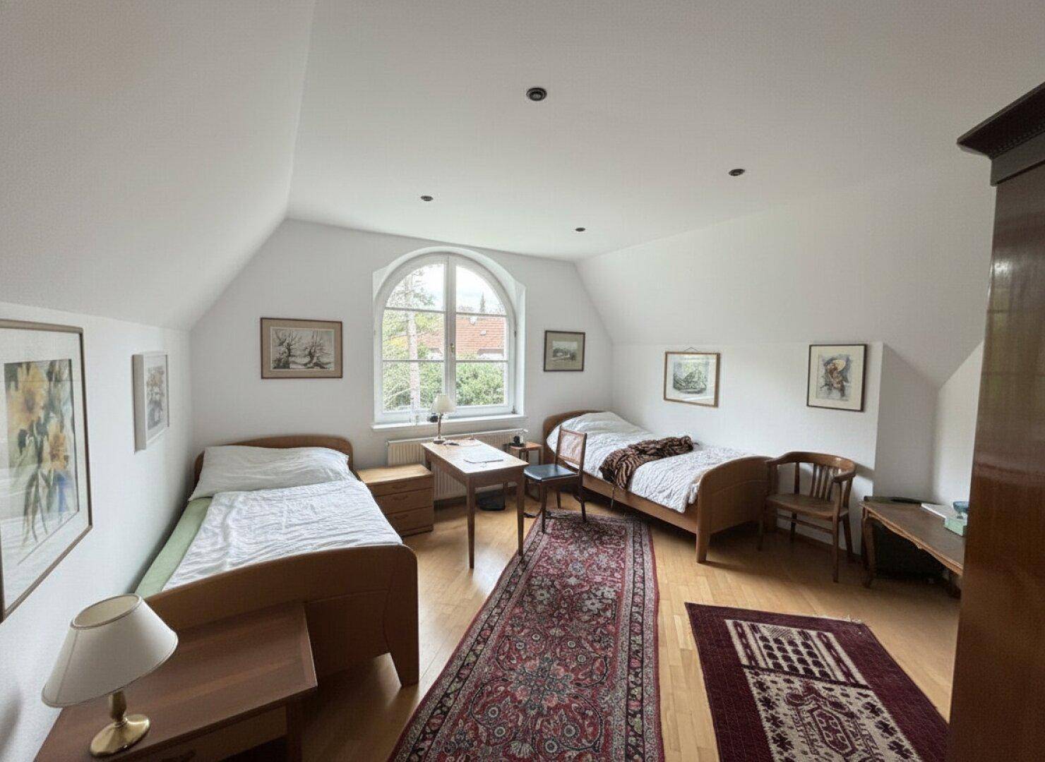 Helles Schlafzimmer im Dachgeschoss mit zwei Betten, Parkettboden und Rundbogenfenster.