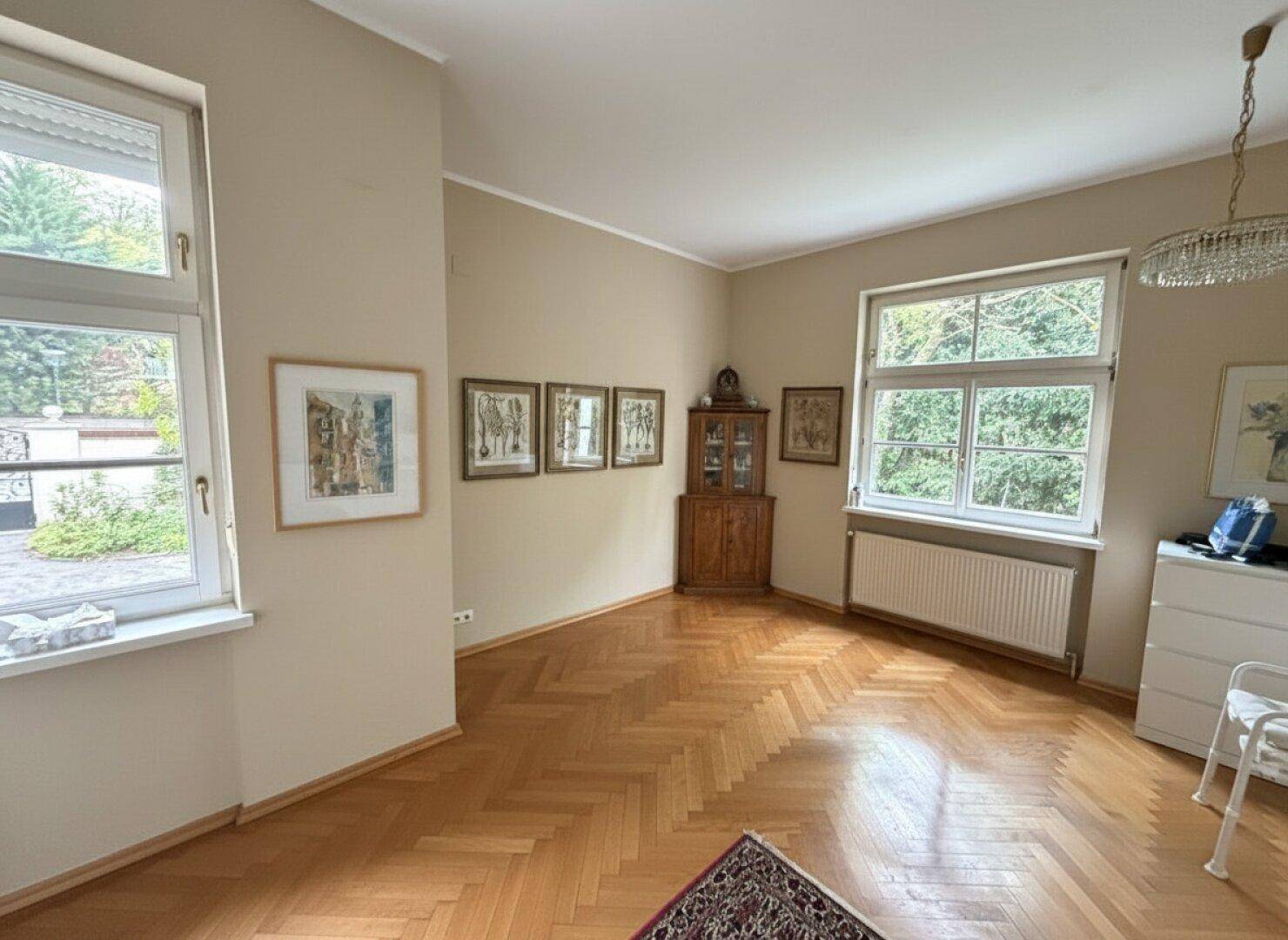 Helles Zimmer mit Parkettboden, großen Fenstern und Blick in den grünen Außenbereich.