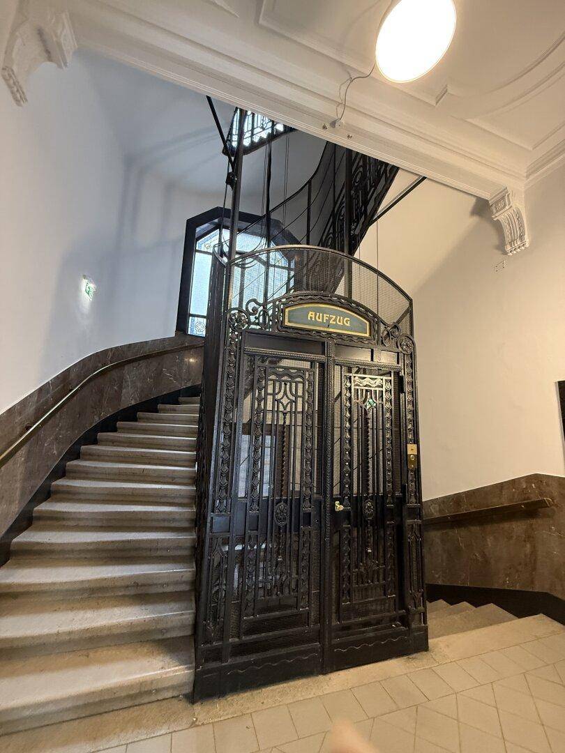 Historischer Gitteraufzug in einem eleganten Treppenhaus mit Marmorstufen und hohen Decken.