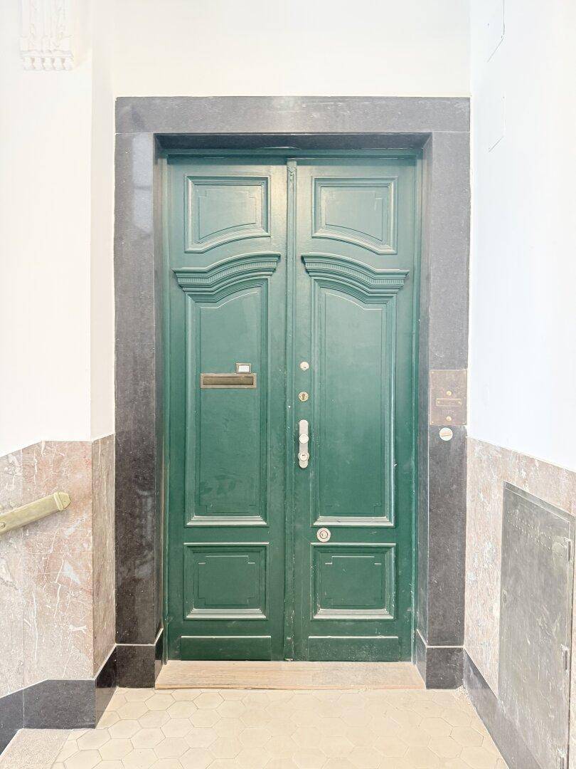 Grüne Eingangstür mit klassischem Design, umrahmt von dunklem Marmor und hellen Wänden.