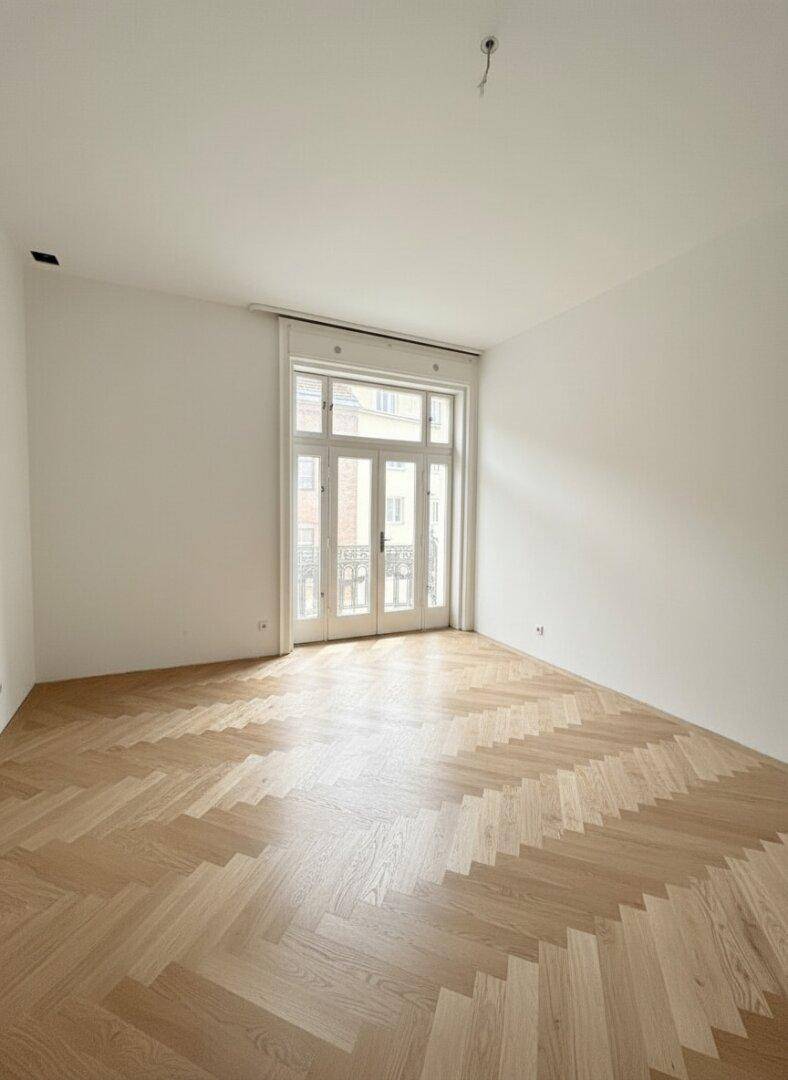 Heller Raum mit Fischgrätparkett und einer französischen Balkontür, die viel Licht hereinlässt.