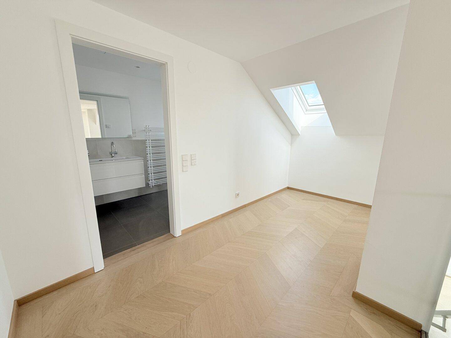 Heller Raum mit Fischgrätparkettboden und Dachfenster, der zu einem modernen Badezimmer führt.