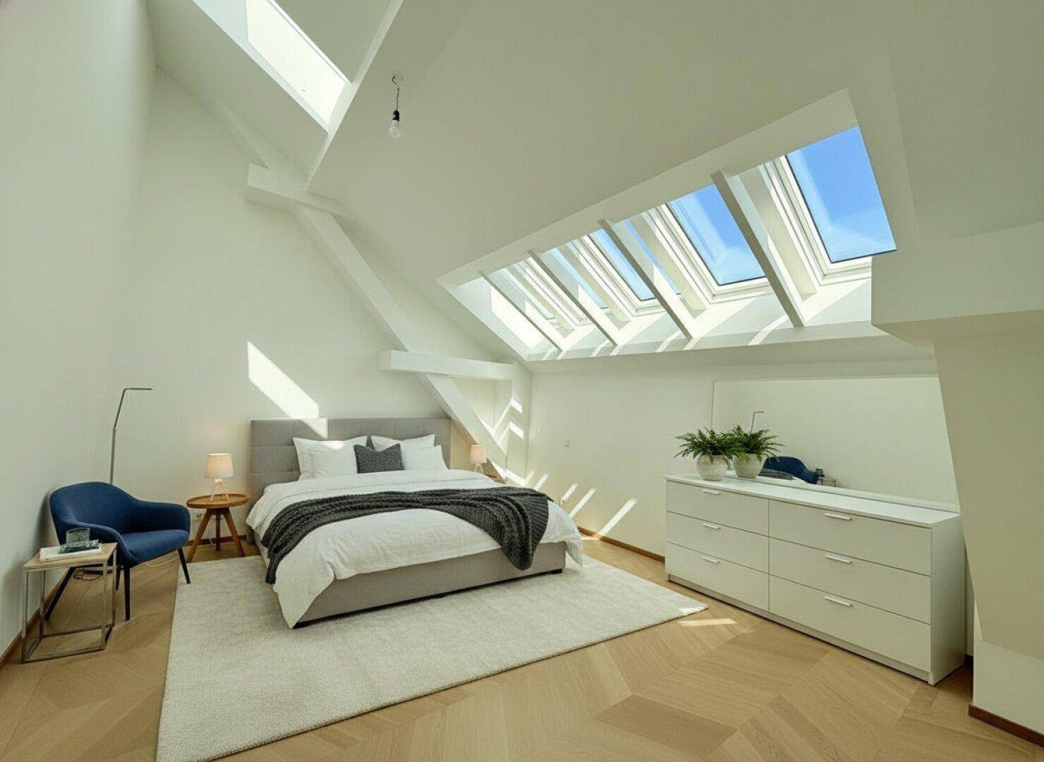Schlafzimmer mit Doppelbett, blauem Sessel und mehreren Dachfenstern, die den Raum mit Tageslicht durchfluten.