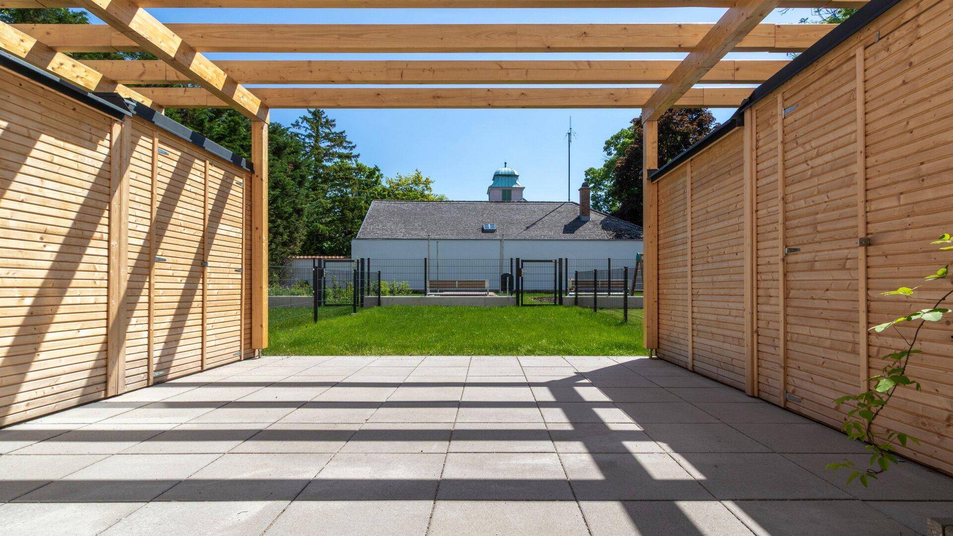 Gepflasterte Terrasse unter einer Holzpergola mit Blick auf eine Grünfläche und ein Gebäude im Hintergrund.