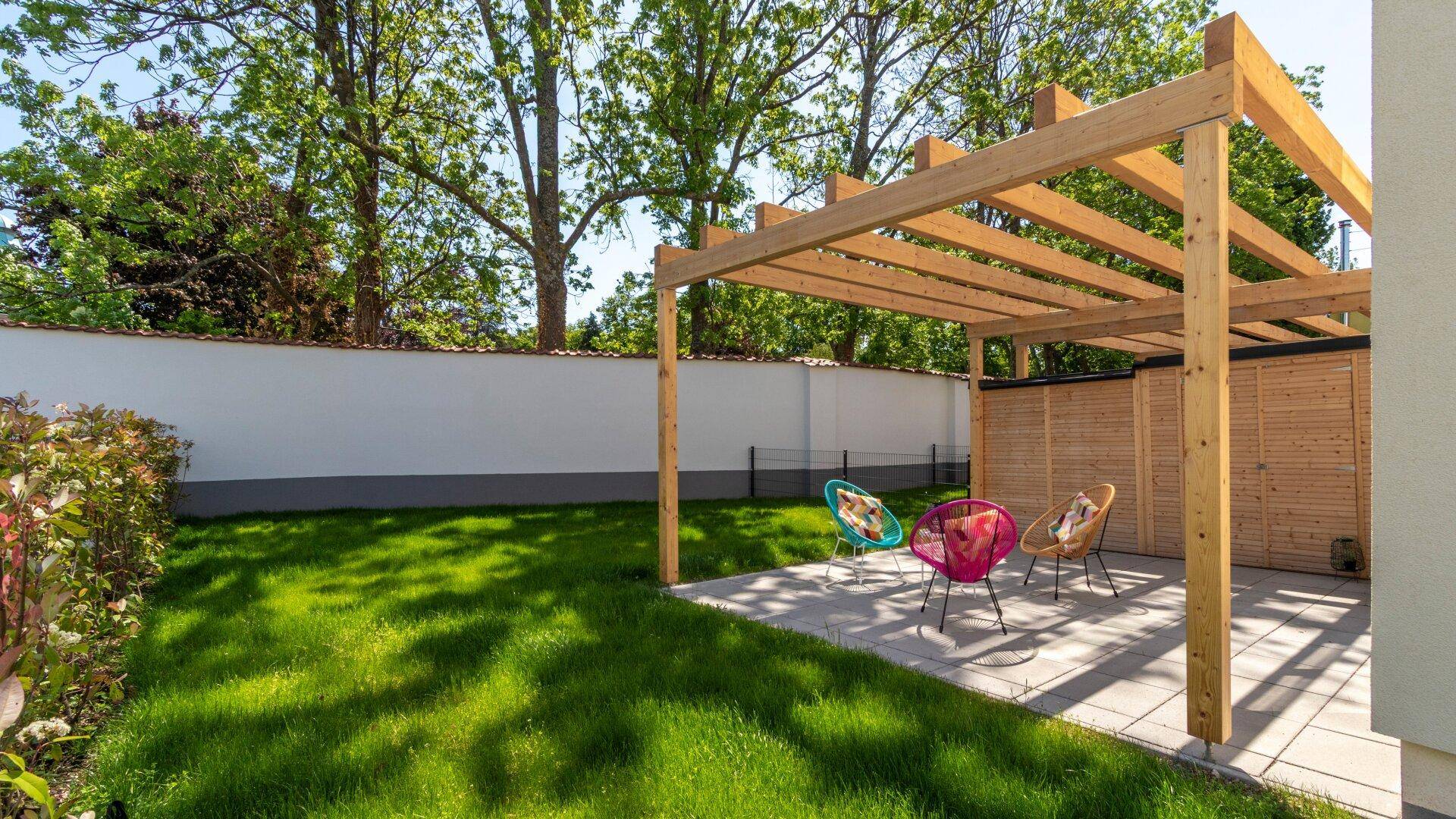 Grüner Gartenbereich mit Rasenfläche und einer gepflasterten Terrasse unter einer Holzpergola mit bunten Stühlen.