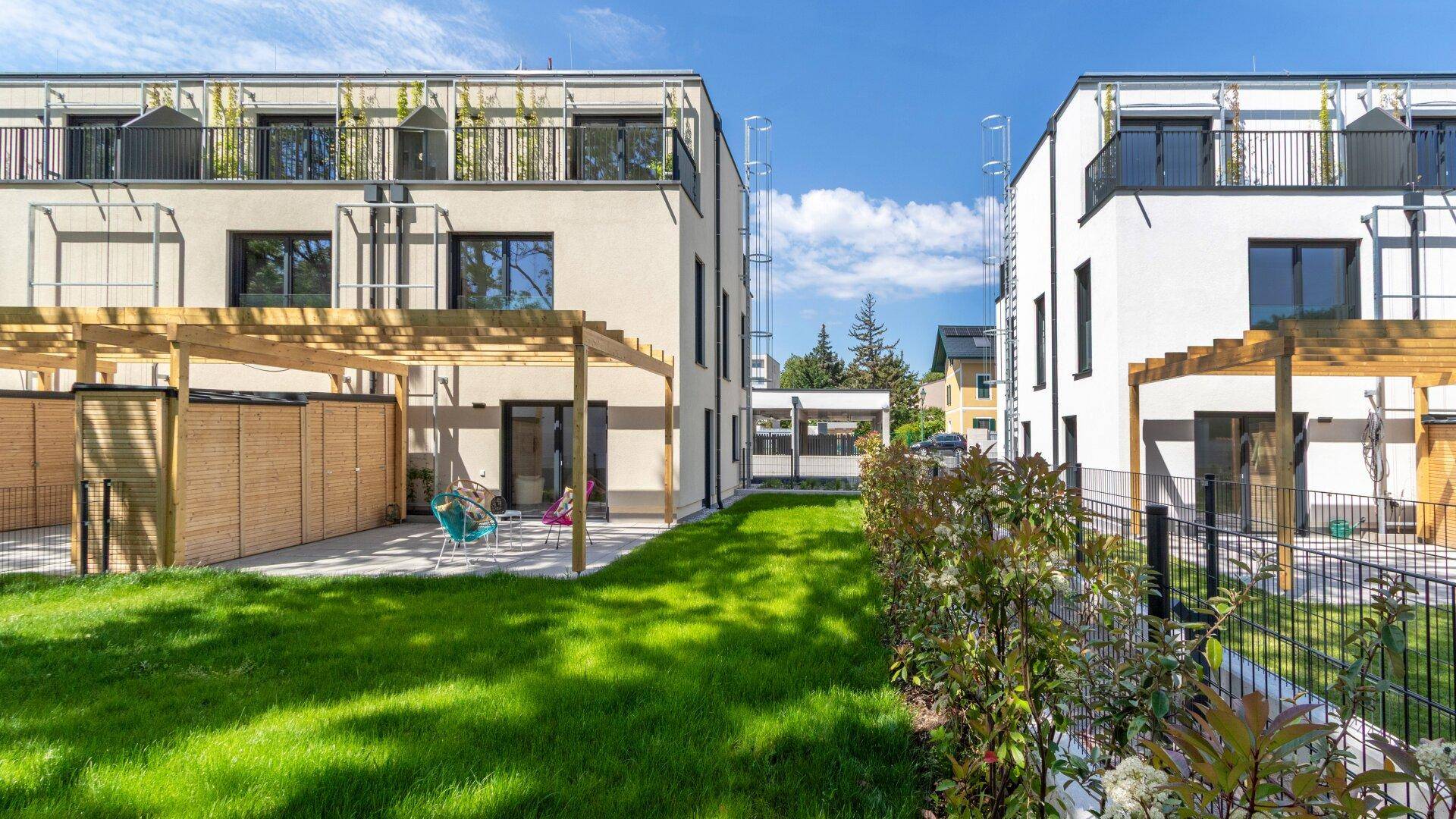 Zwei moderne Wohngebäude mit privaten Gärten, Terrassen und Holzpergolen in sonniger Umgebung.