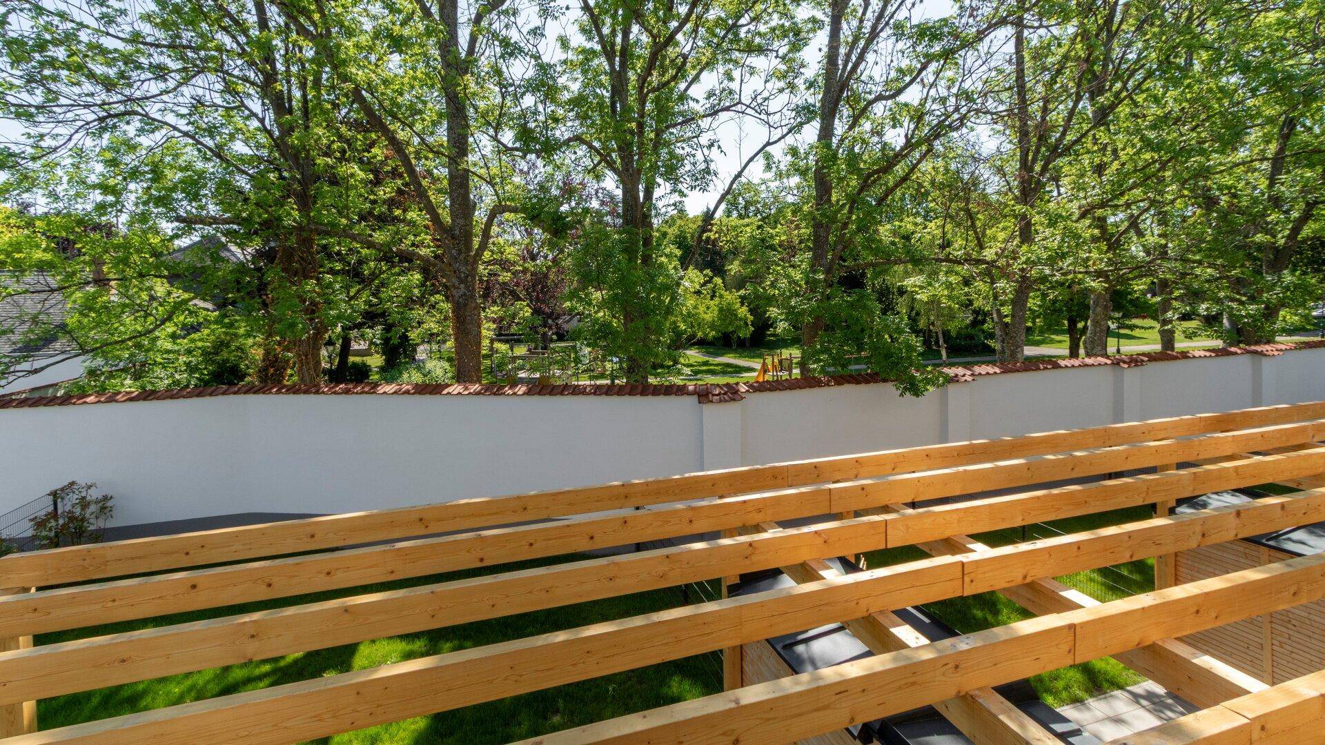 Blick über eine Holzpergola auf eine weiße Mauer und alte Bäume, mit einem Spielplatz im Hintergrund.