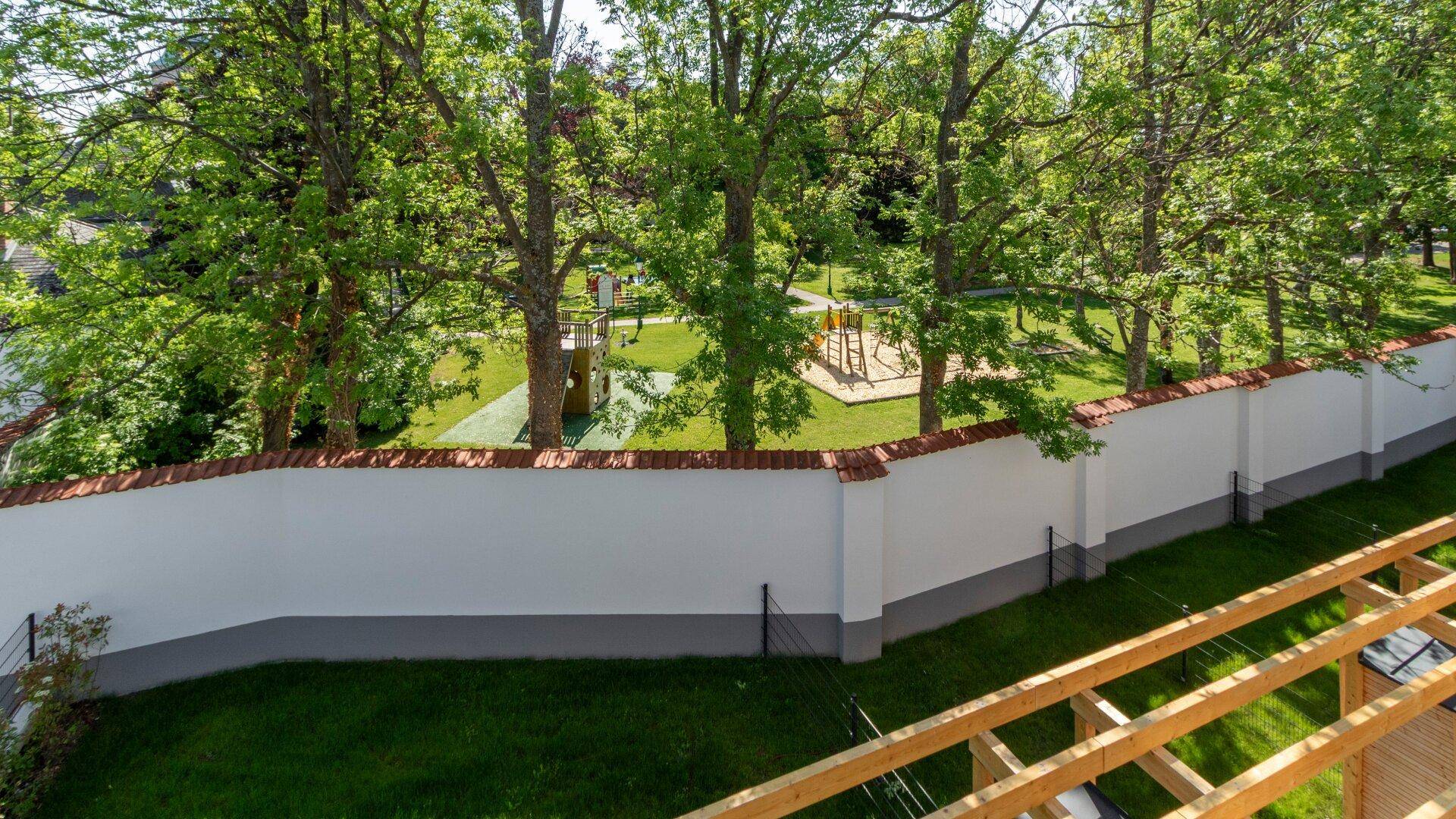 Blick über eine weiße Mauer auf einen Spielplatz mit Bäumen und viel Grün unter blauem Himmel.