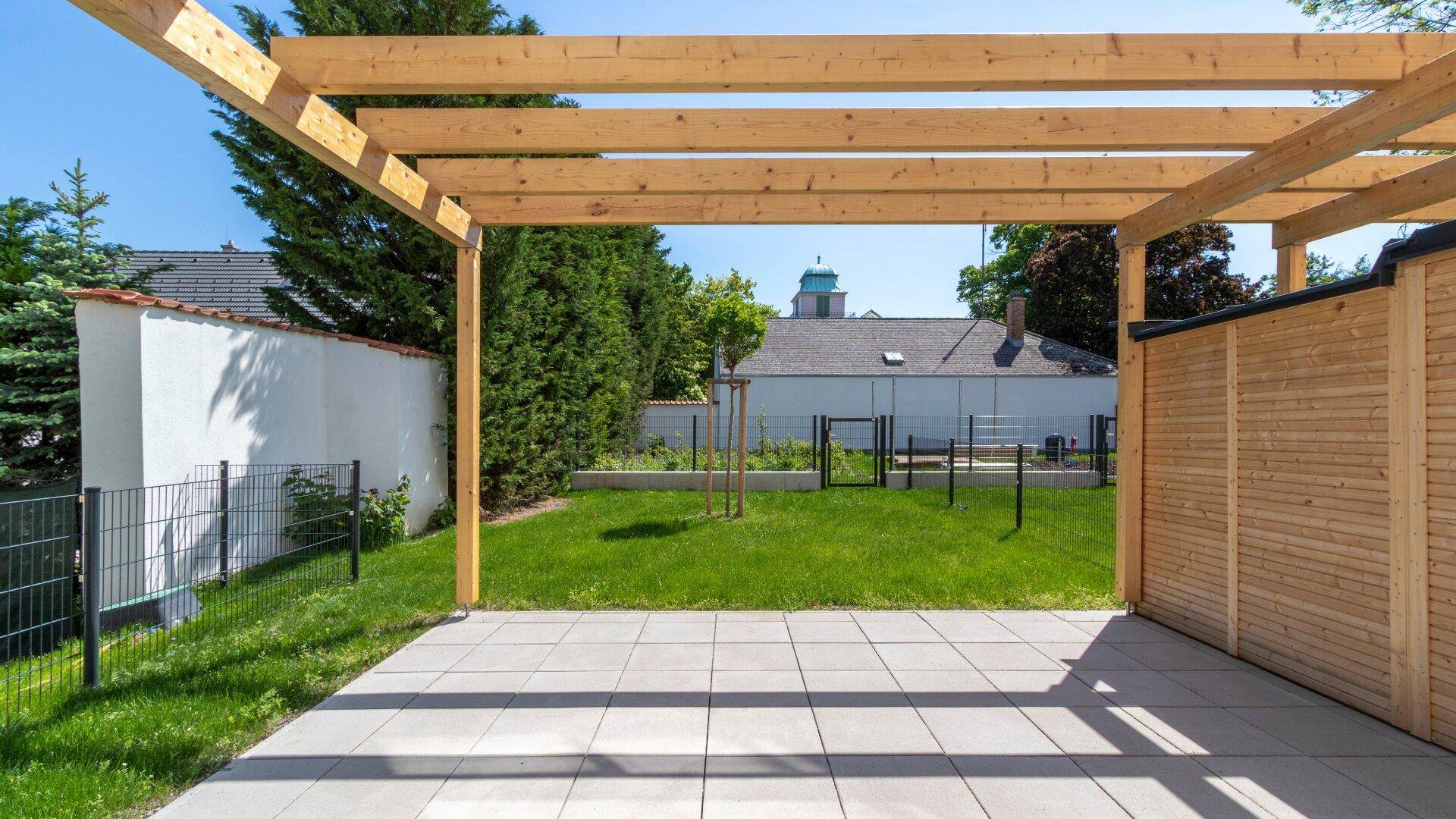 Sonniger Terrassenbereich mit Holzpergola und Fliesenboden, angrenzend an eine grüne Rasenfläche.