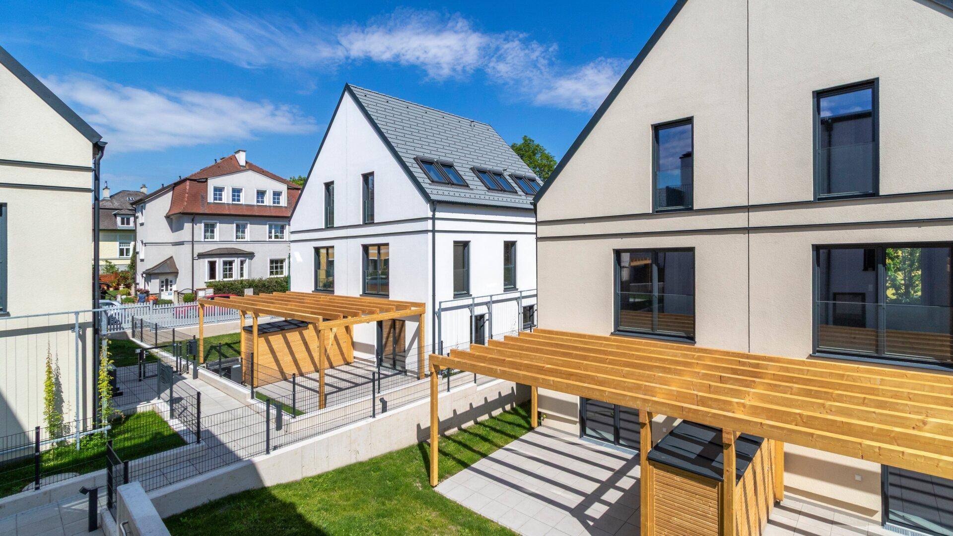 Moderne Reihenhäuser mit Satteldächern und Pergolen im Vorgartenbereich unter blauem Himmel.