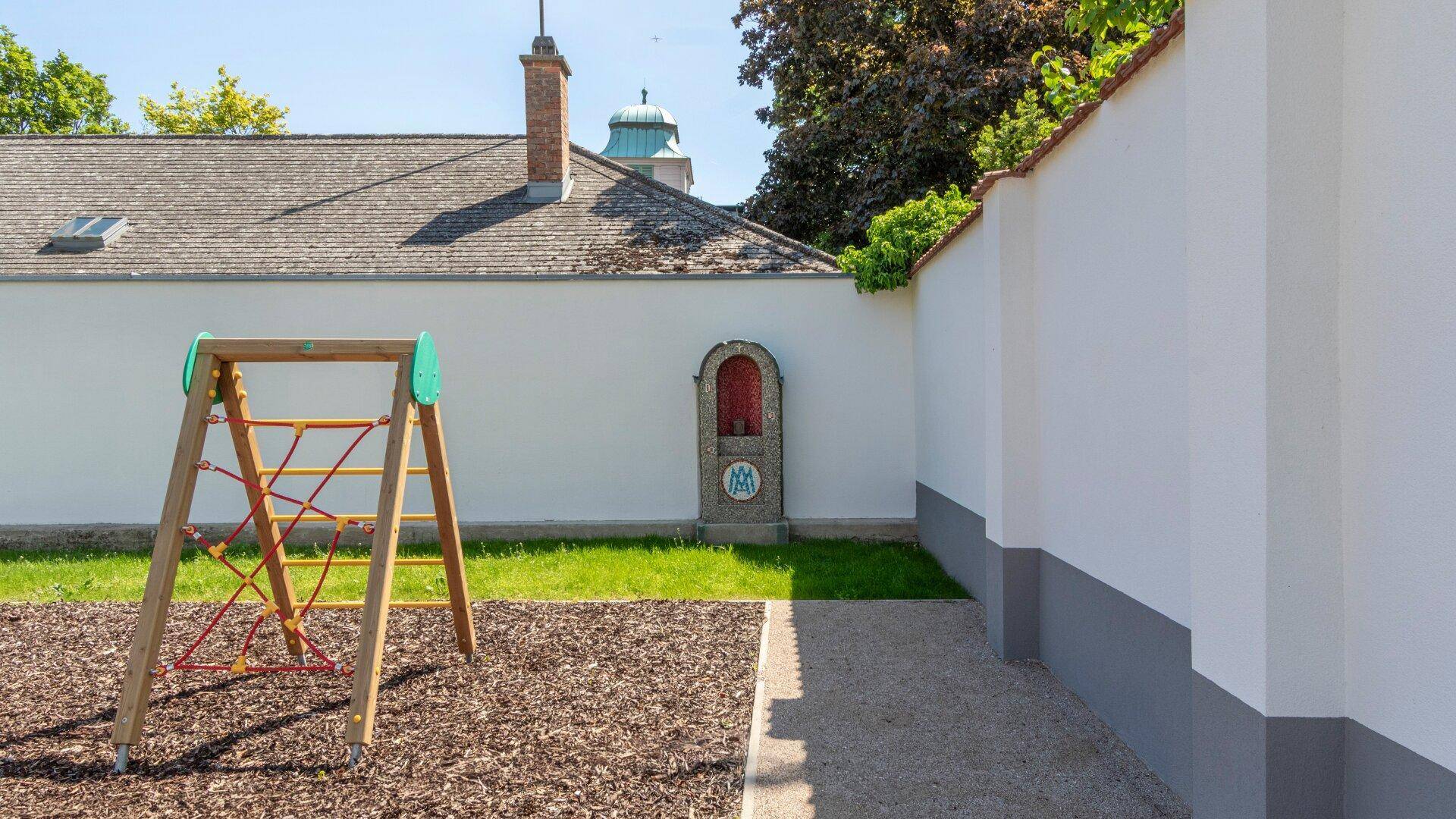Spielplatz mit Klettergerüst und Sandbereich, angrenzend an eine weiße Mauer mit kleinem Schrein.