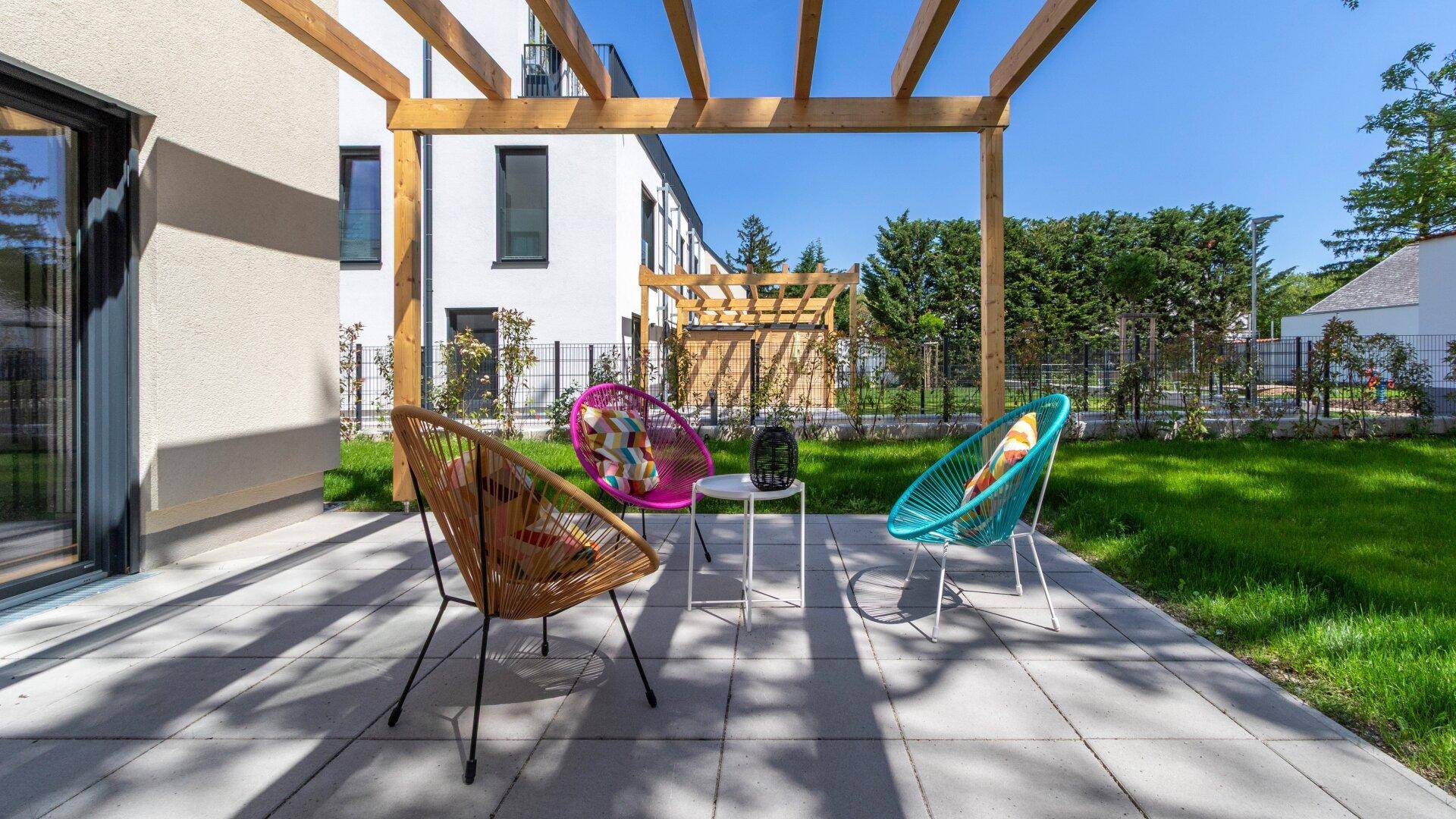 Sonniger Terrassenbereich mit bunten Stühlen unter einer Holzpergola, umgeben von Rasen.