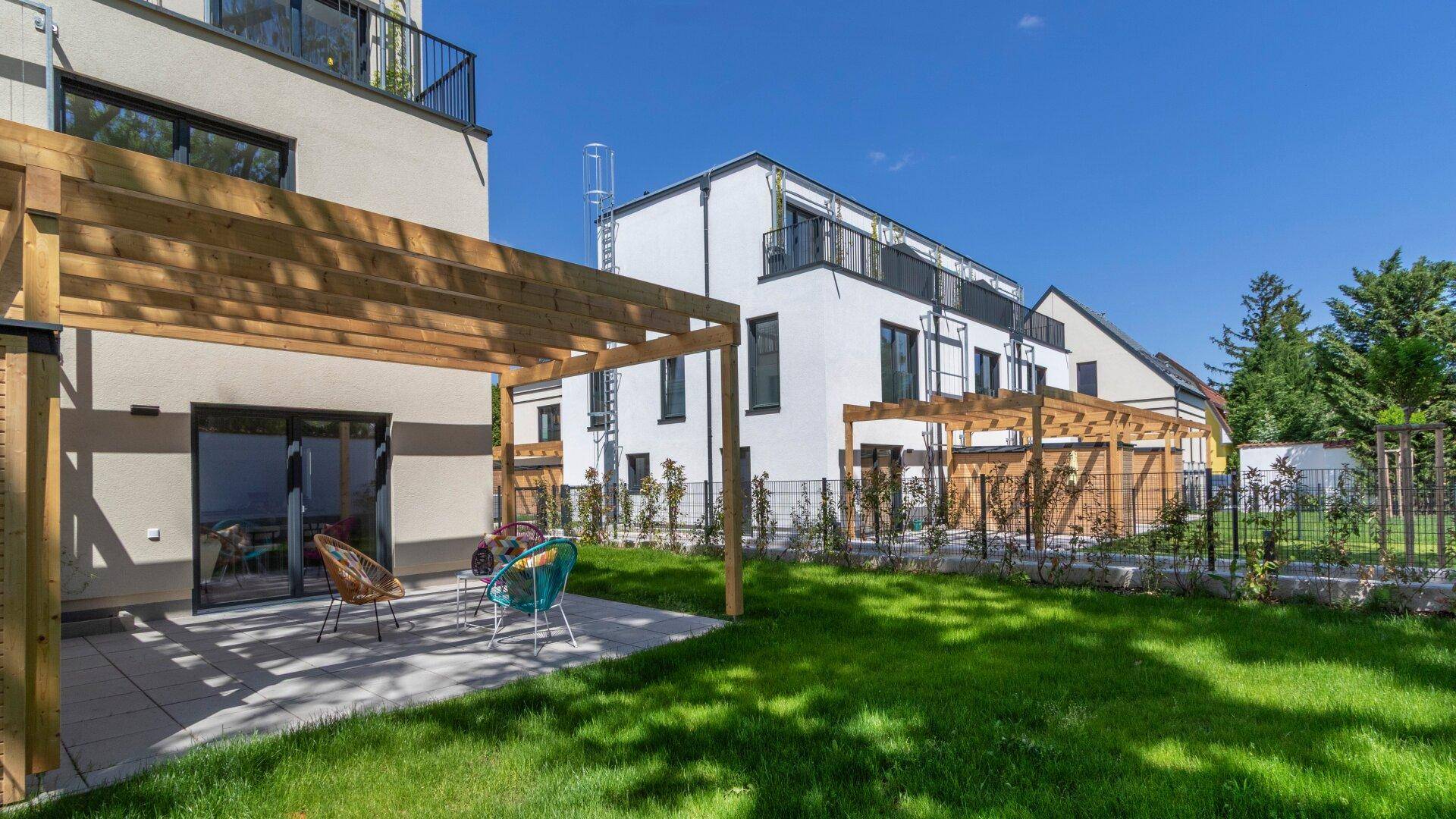 Außenansicht eines modernen Neubaus mit sonniger Terrasse, Pergola und gepflegtem Garten.