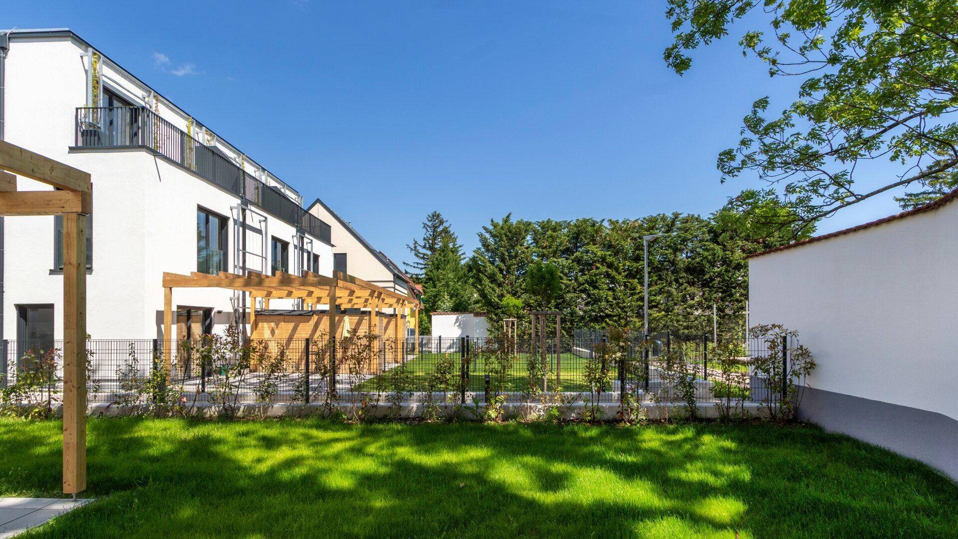 Moderne Gebäudefassade mit Gartenfläche, Zaun und hohen Hecken im Hintergrund.
