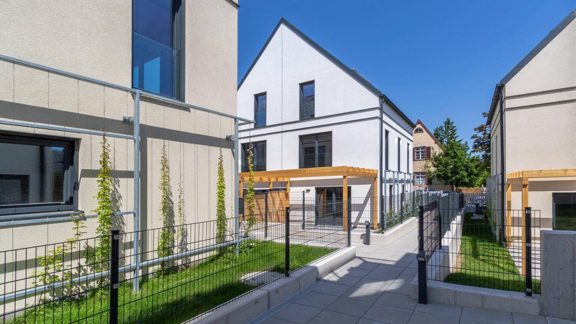 Gepflasterter Weg zwischen zwei modernen Reihenhäusern mit kleinen Grünflächen und Pergolen.