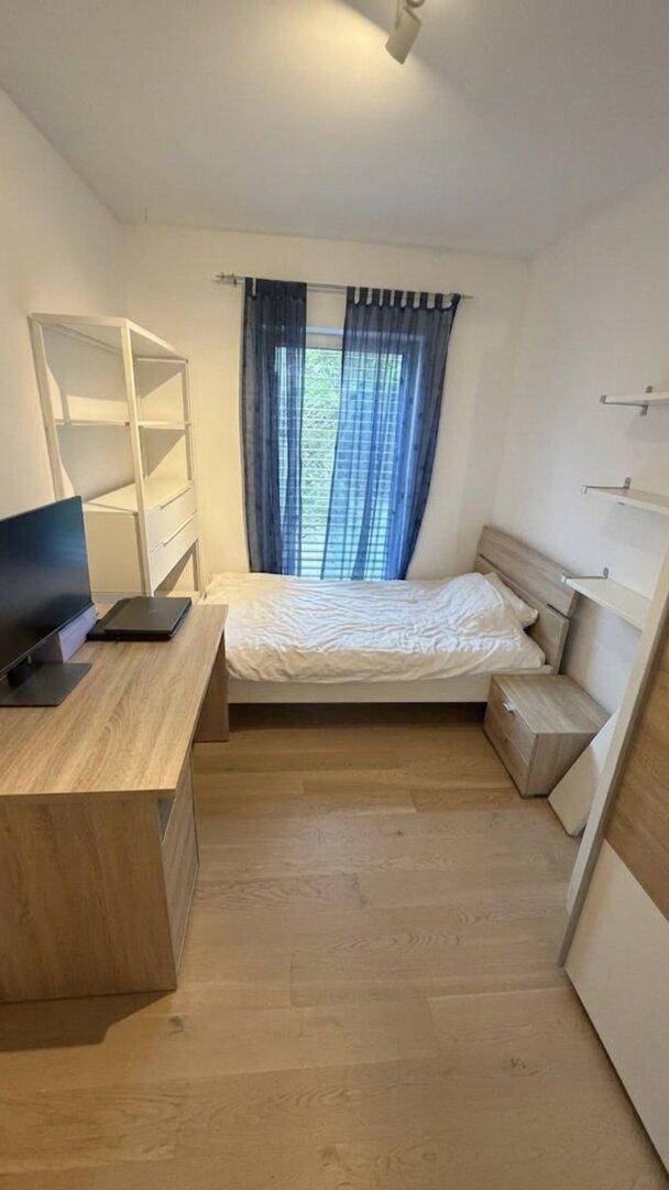 Gemütliches Schlafzimmer mit Holzboden, Einzelbett, Schreibtisch und blauen Vorhängen am Fenster.