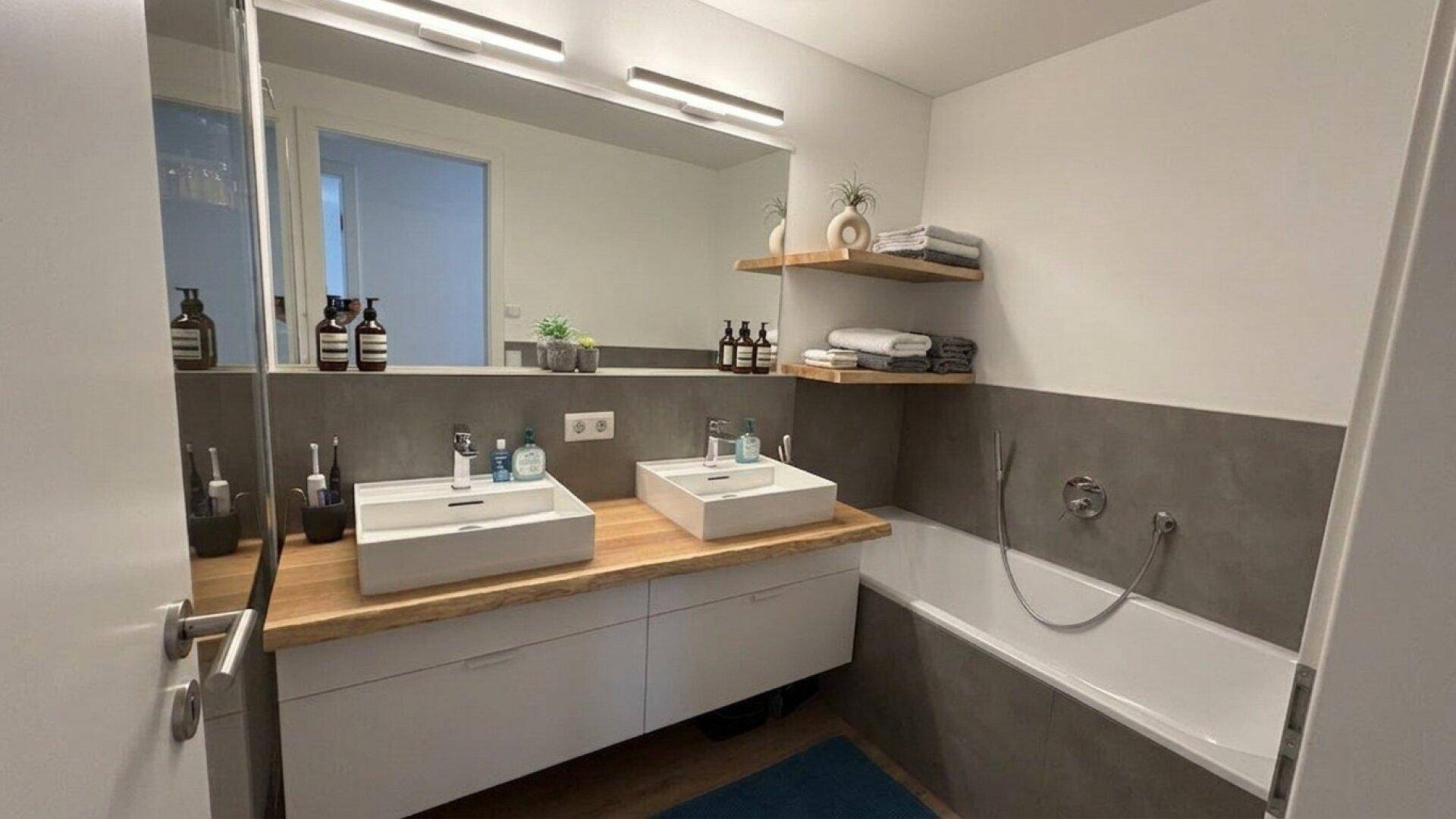 Modernes Badezimmer mit Doppelwaschbecken, Badewanne und Holzregalen für Handtücher.