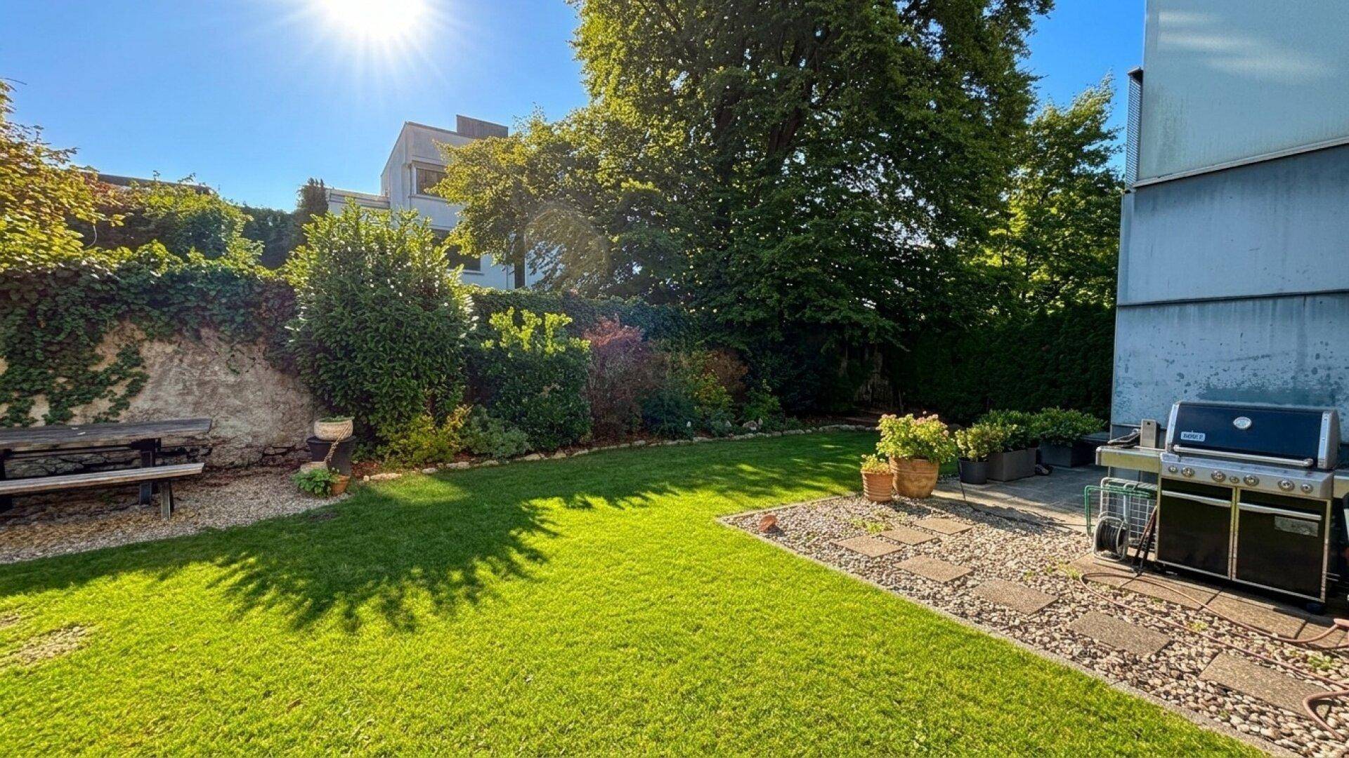 Großer, sonniger Garten mit Rasenfläche, alten Bäumen und einem Grillplatz im Freien.