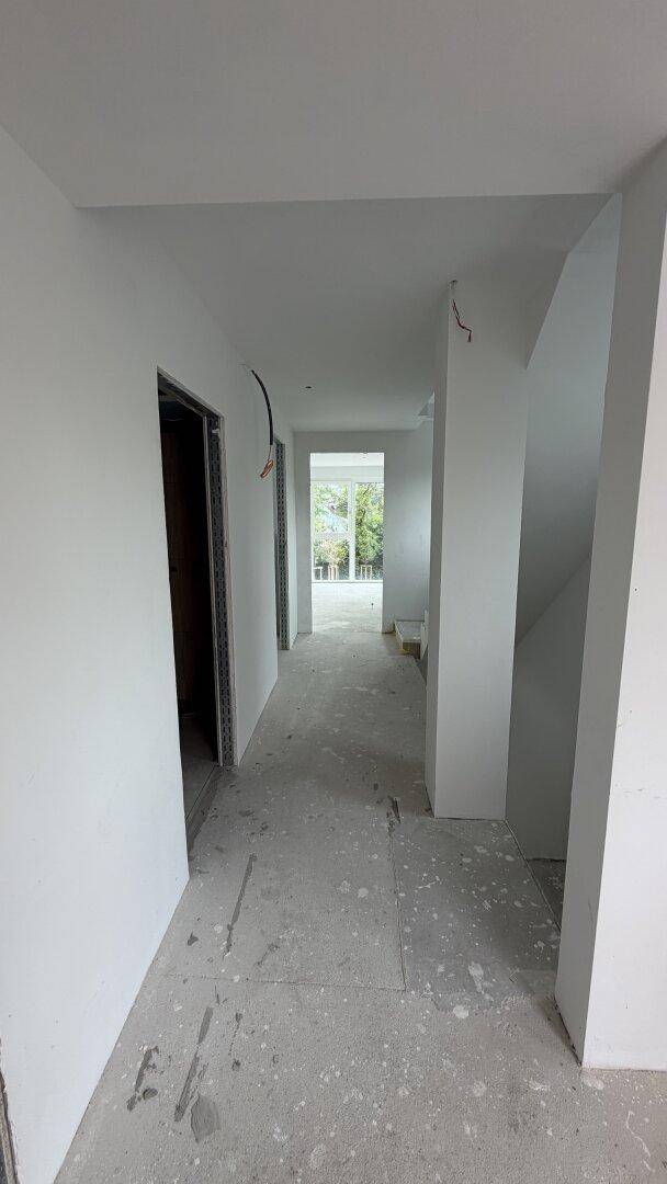 Unfertiger Flur im Neubau mit Betonboden und weißen Wänden, Blick ins Grüne durch ein Fenster.