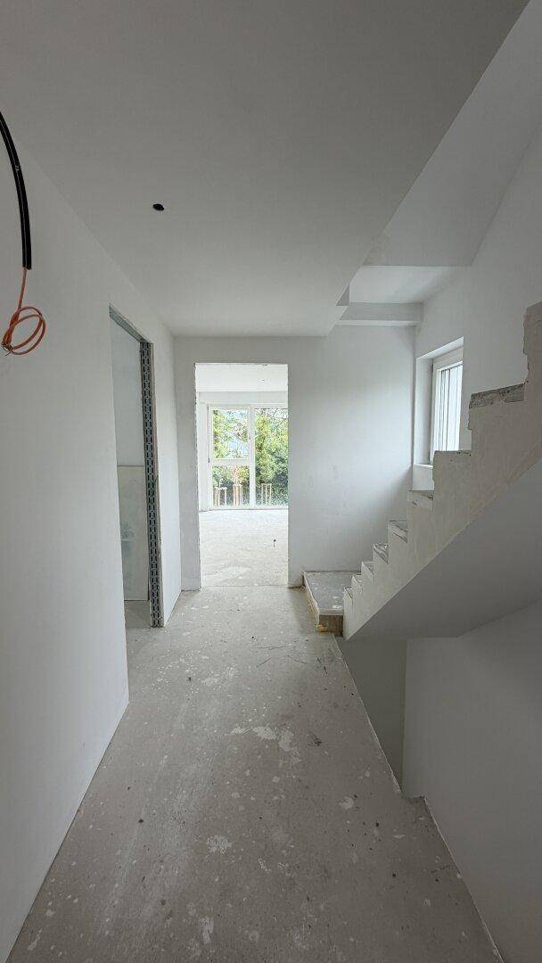 Flur im Rohbau mit Treppenaufgang und Betonboden, helle Wände und Blick nach draußen.