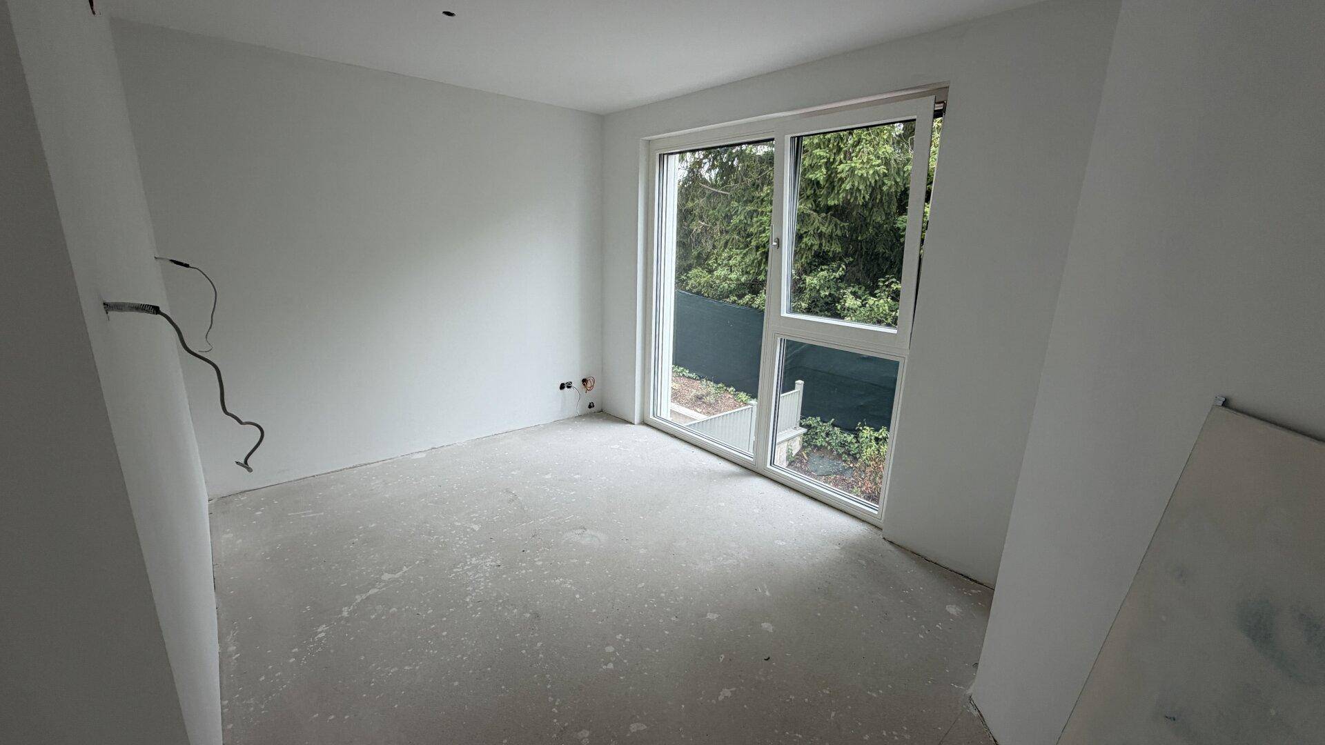 Helles Zimmer im Rohbau mit Betonboden und großem Fenster, das Zugang zum Garten bietet.