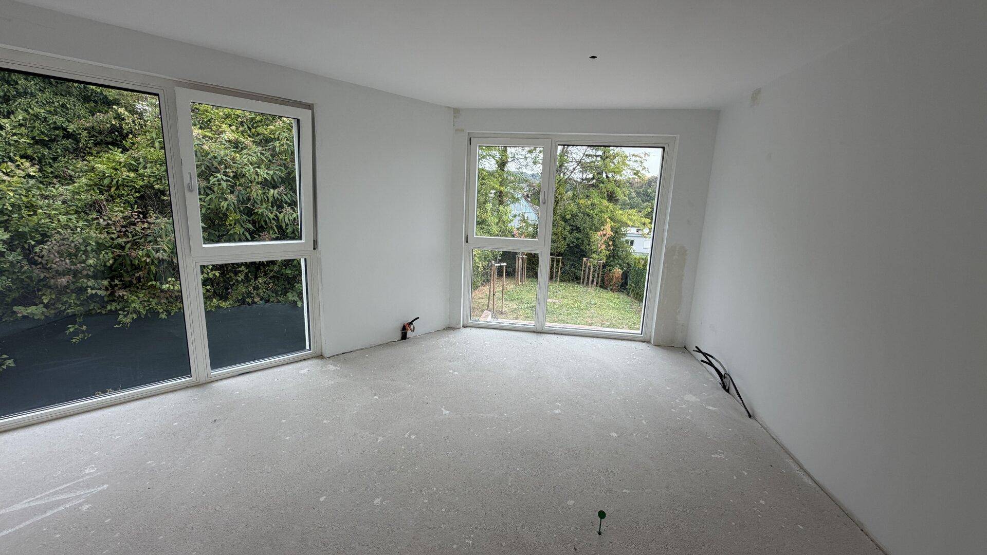 Leeres Schlafzimmer im Rohbau mit großen Fenstern und Blick auf den zukünftigen Garten.