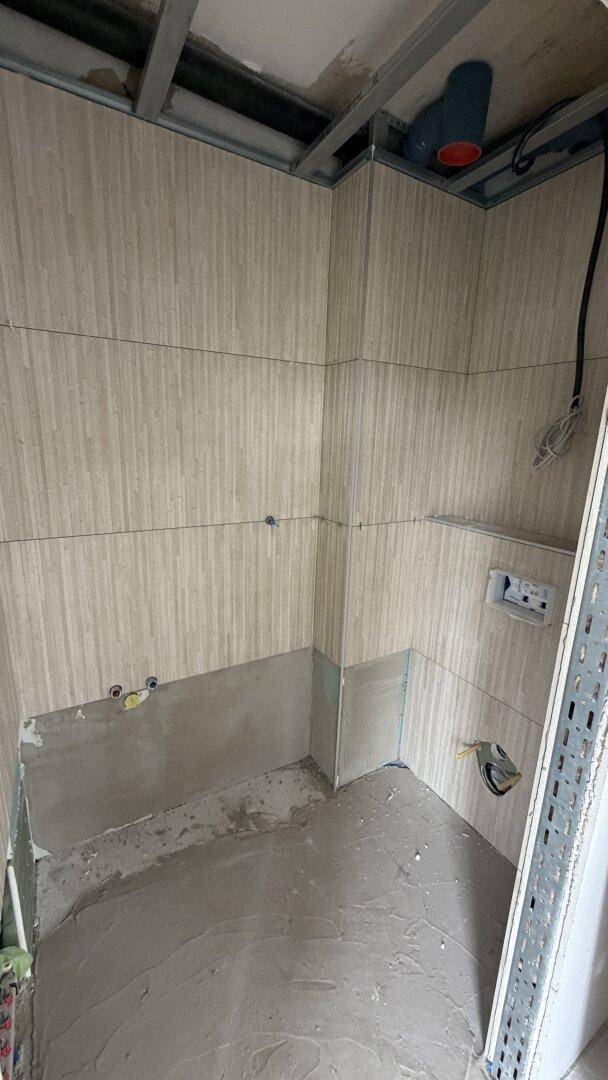 Badezimmer im Rohbau mit vorbereiteten Anschlüssen für Dusche und WC, geflieste Wände.