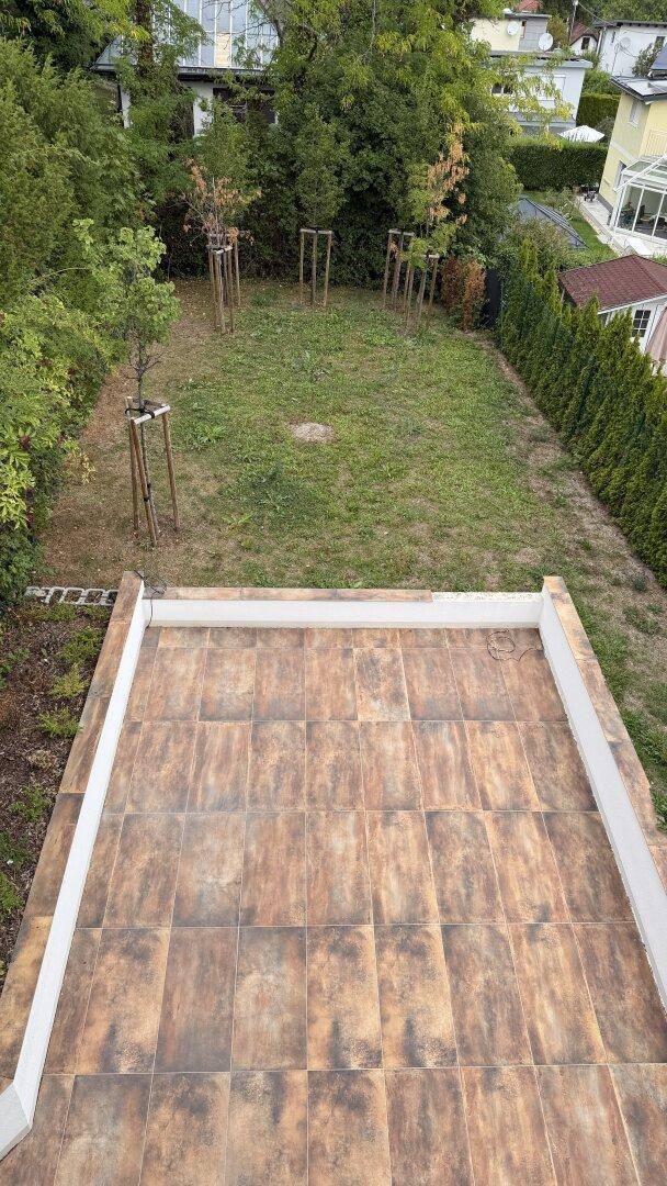 Überblick über die geflieste Terrasse und den Gartenbereich mit Rasen und Hecken, von oben gesehen.