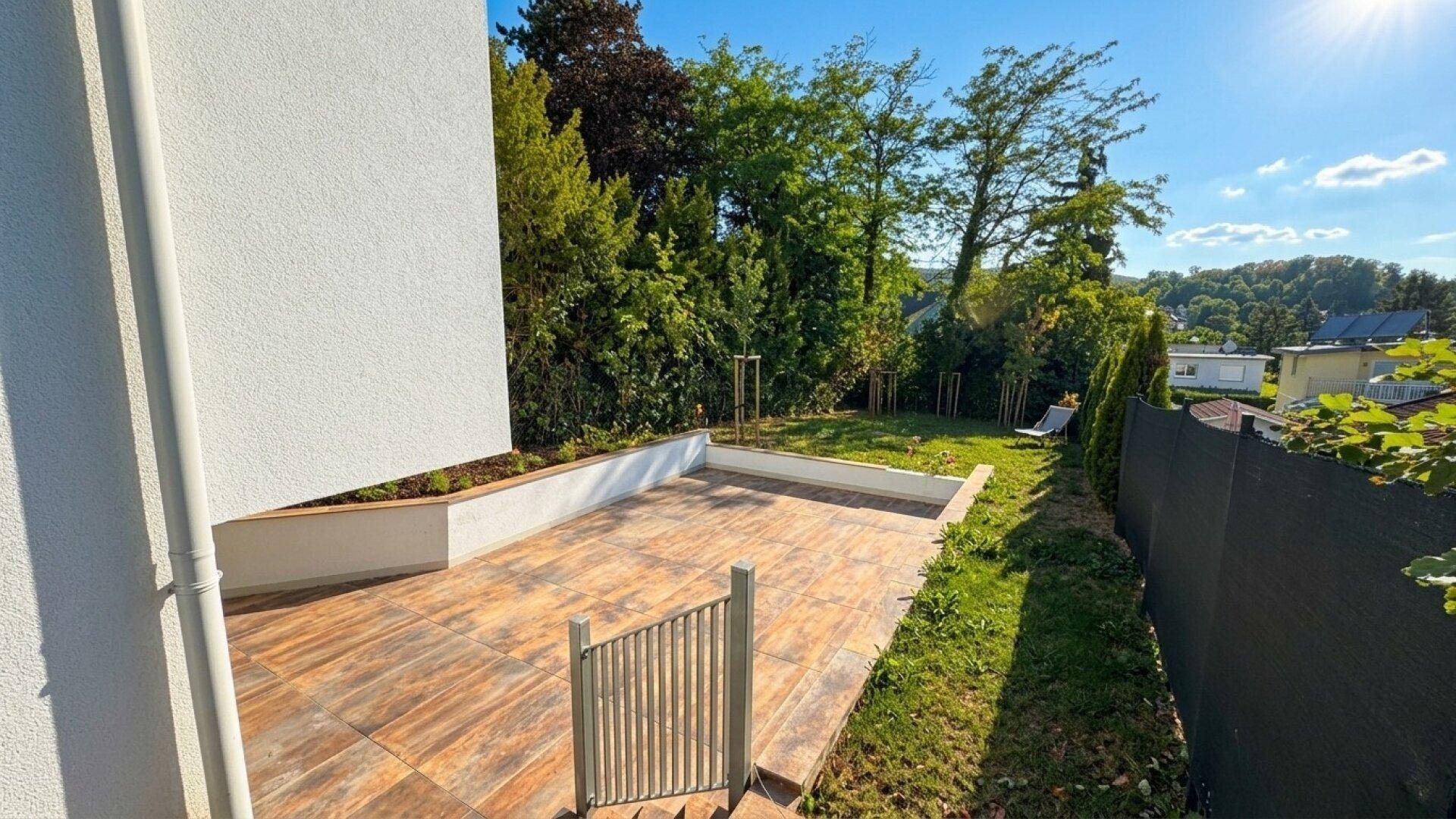 Hinterer Gartenbereich mit gepflasterter Terrasse und Rasenfläche, umgeben von Bäumen.