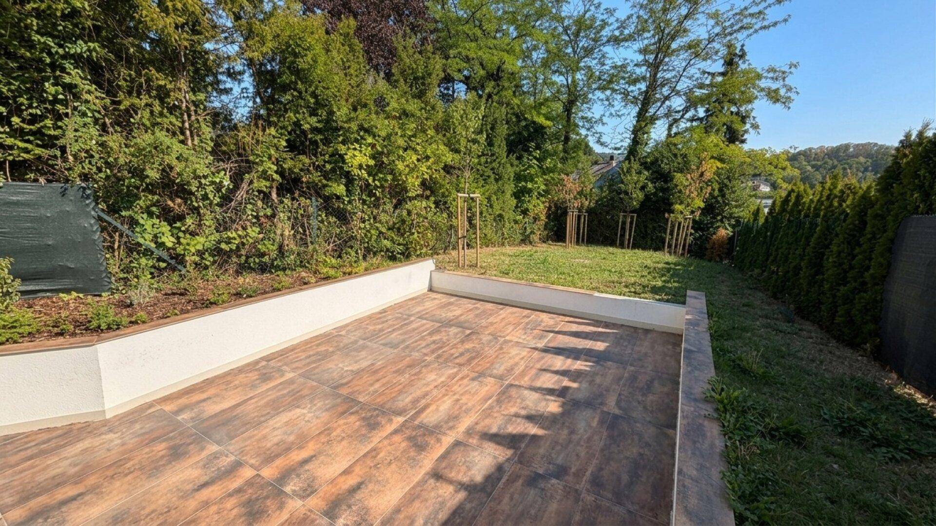 Große geflieste Terrasse mit erhöhten Blumenbeeten und angrenzendem Rasenbereich, umgeben von viel Grün.