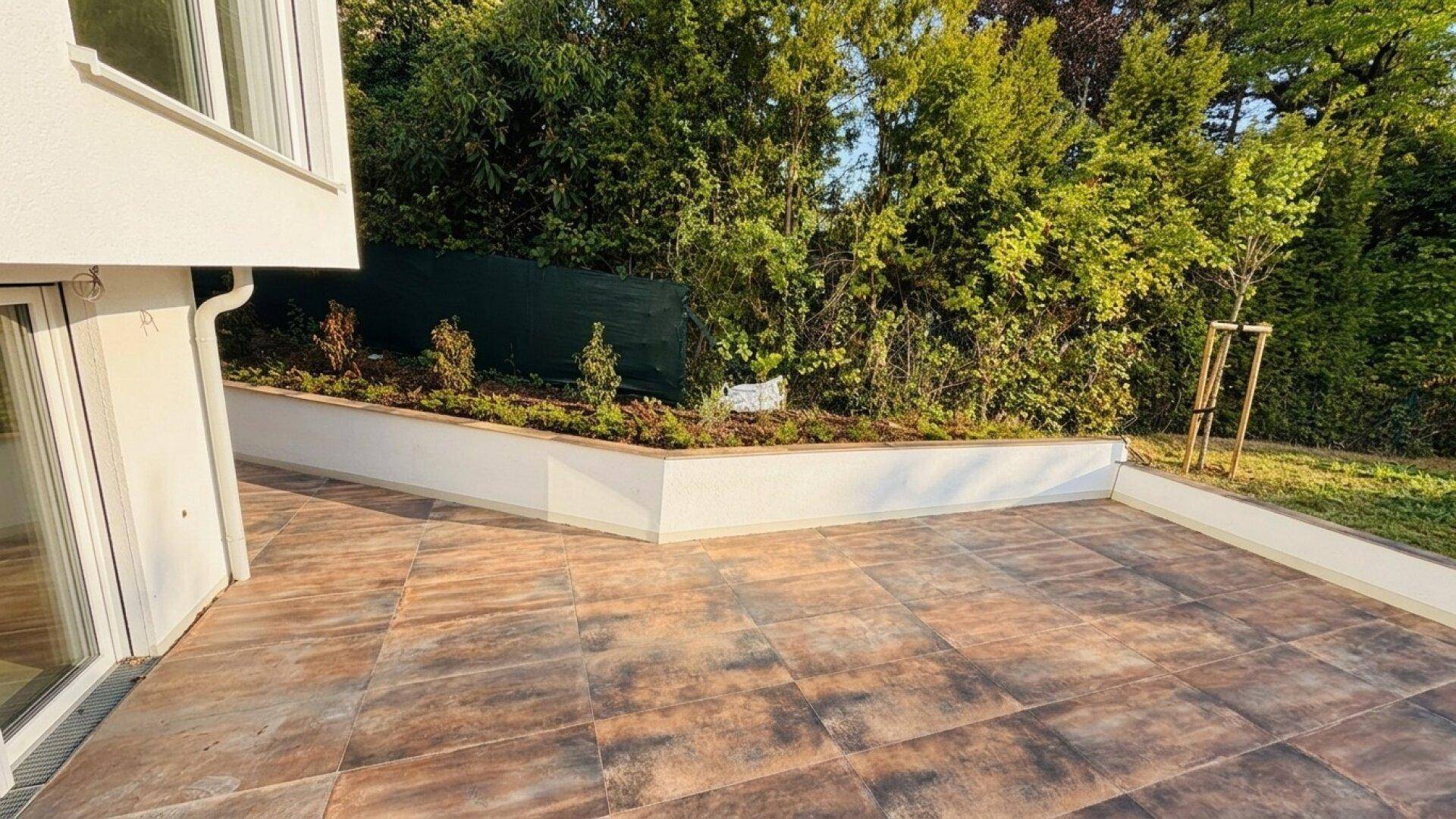Sonniger Terrassenbereich am Haus mit gefliestem Boden und bepflanzten Hochbeeten, umgeben von Bäumen.