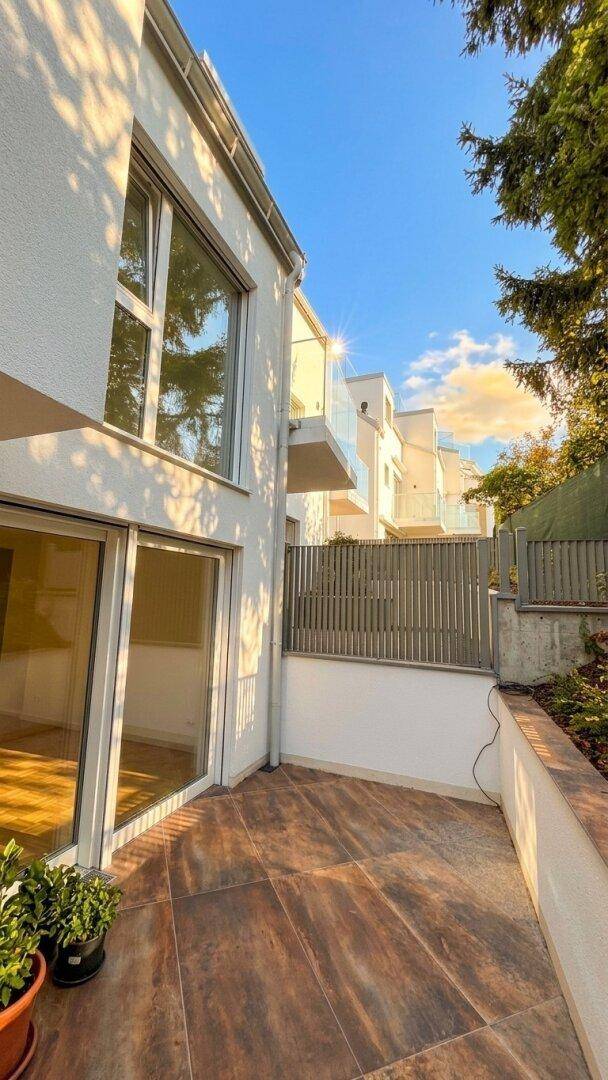 Sonniger Patio-Bereich am modernen Gebäude mit gefliestem Boden und Blick auf weitere Neubauten.