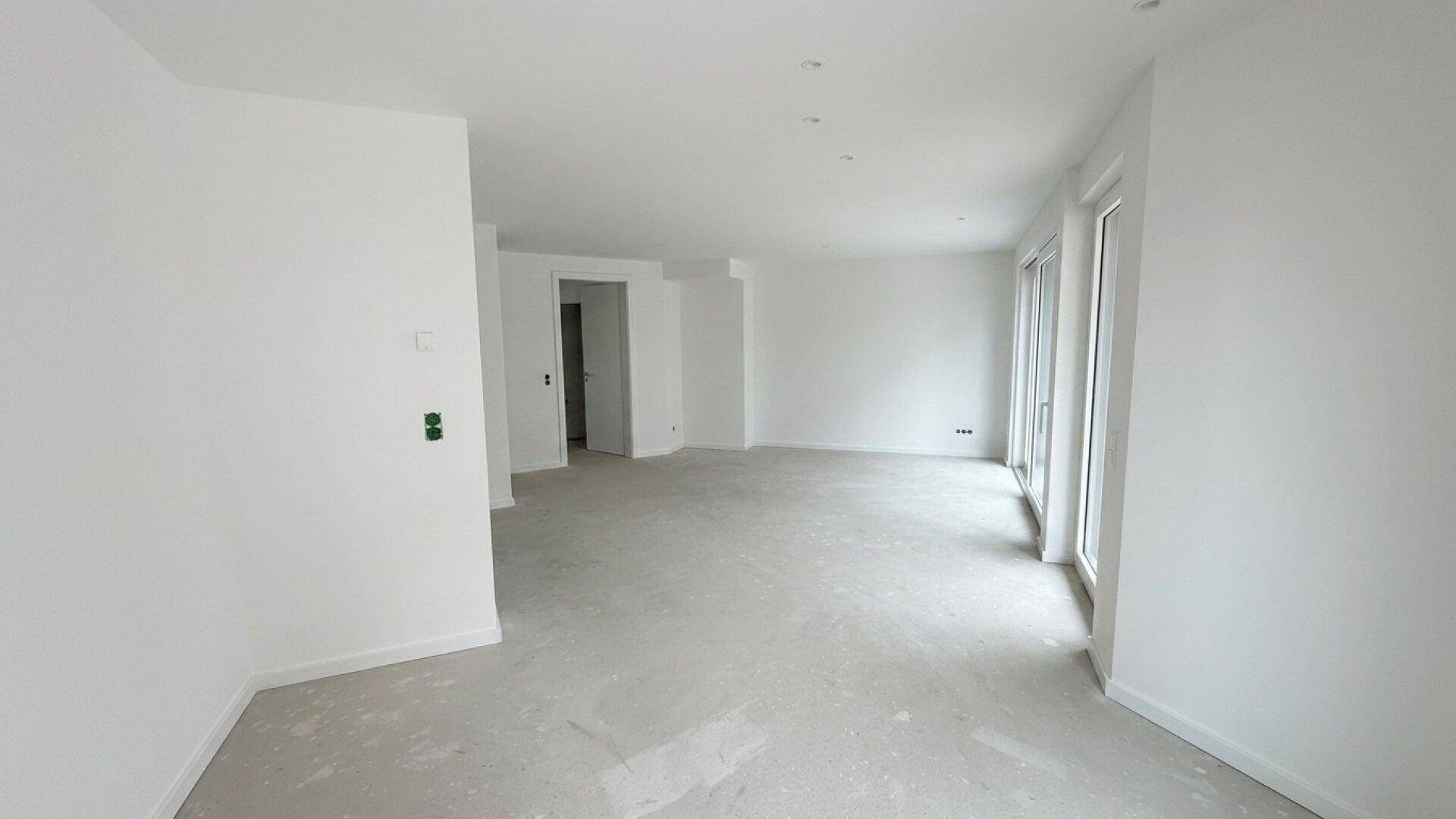 Leerer Raum im Rohbauzustand mit weißen Wänden und viel Tageslicht durch große Fenster.