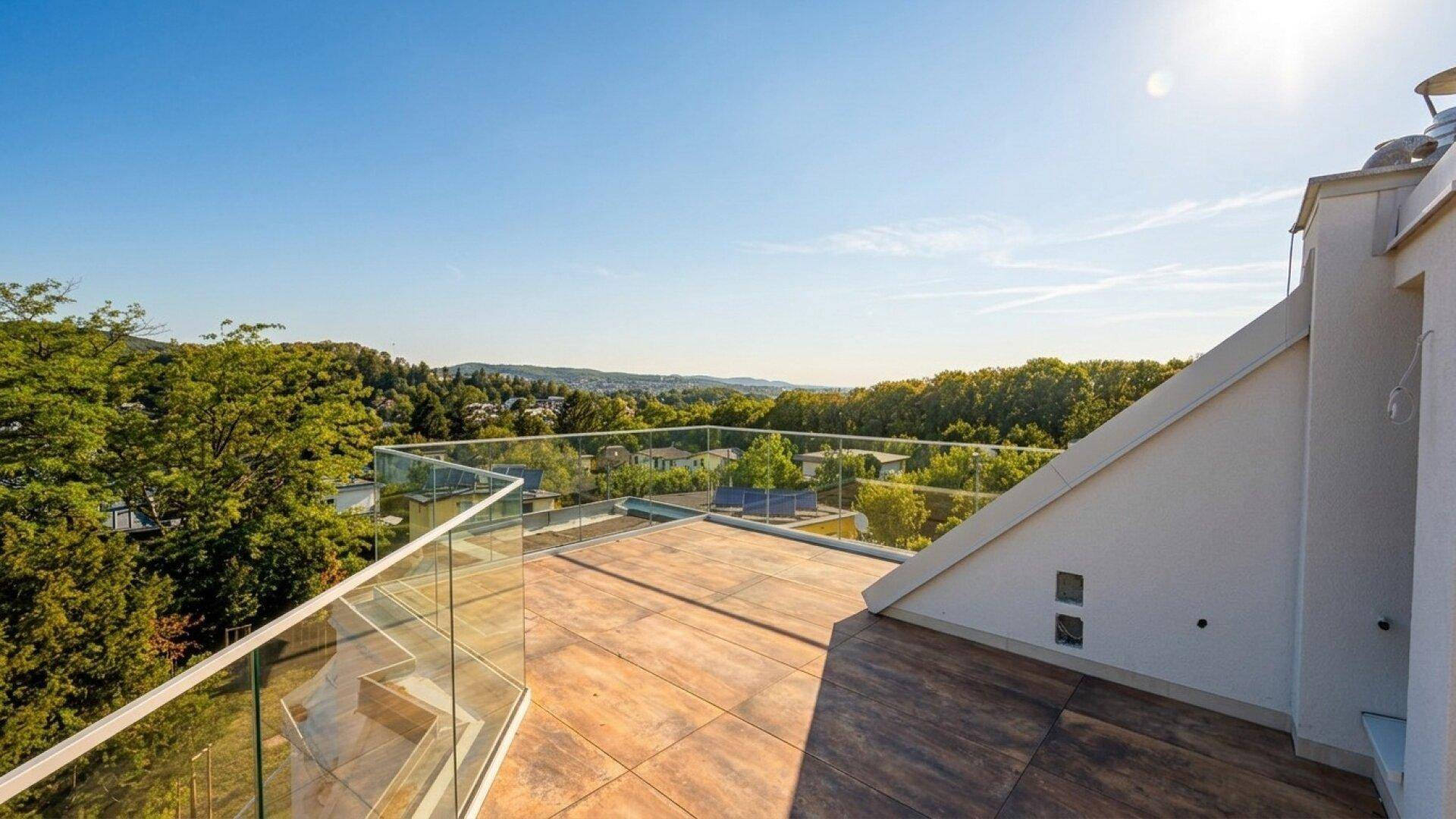 Helle Dachterrasse mit Glasgeländer und weitem Blick über die grüne Umgebung unter blauem Himmel.