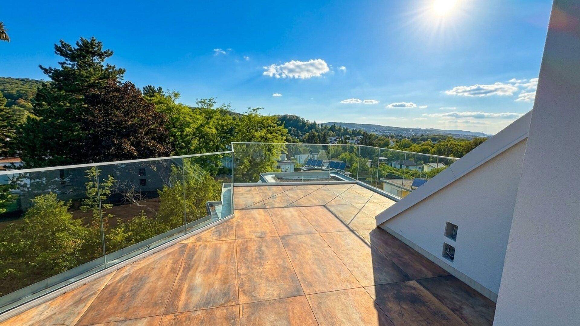 Sonniger Dachterrassenbereich mit Glasgeländer und weitem Blick über die grüne Landschaft und Häuser.