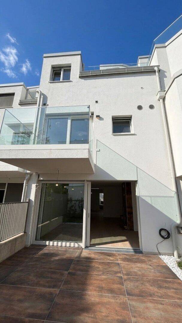 Rückseite des Gebäudes mit ebenerdiger Terrasse und darüberliegendem Balkon mit Glasgeländer.