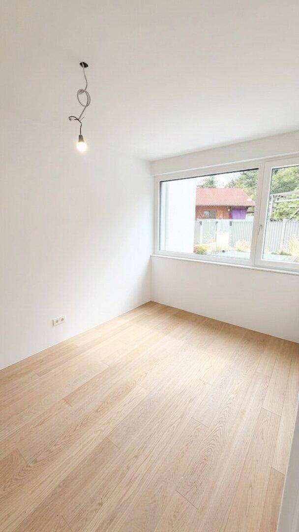 Helles, leeres Zimmer mit Holzfußboden und großem Fenster, ideal als Schlafzimmer.