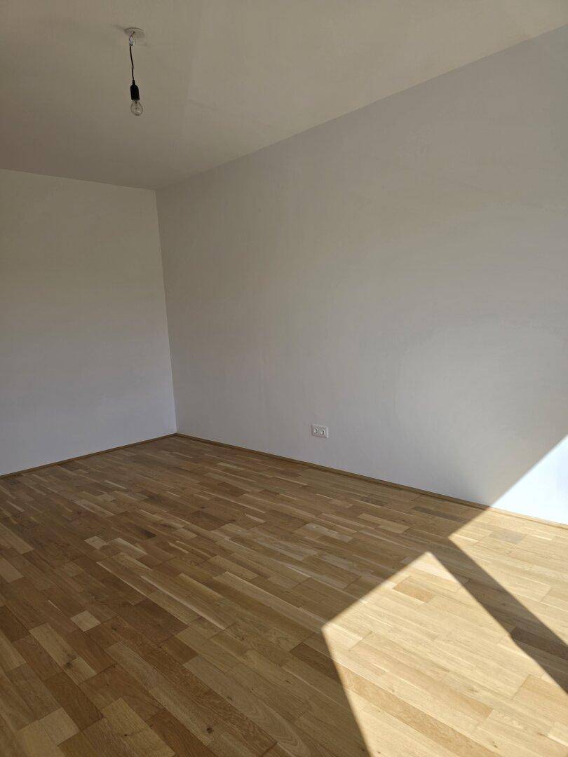 Leerer Raum mit hellem Holzfußboden und weißen Wänden, viel Tageslicht durchflutet.