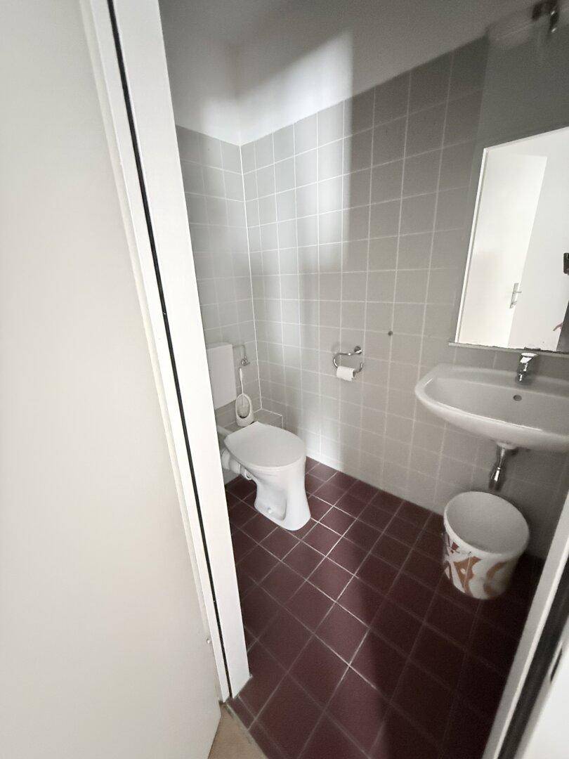 Kleines Gäste-WC mit grauen Wandfliesen, roten Bodenfliesen und einem Waschbecken mit Spiegel.