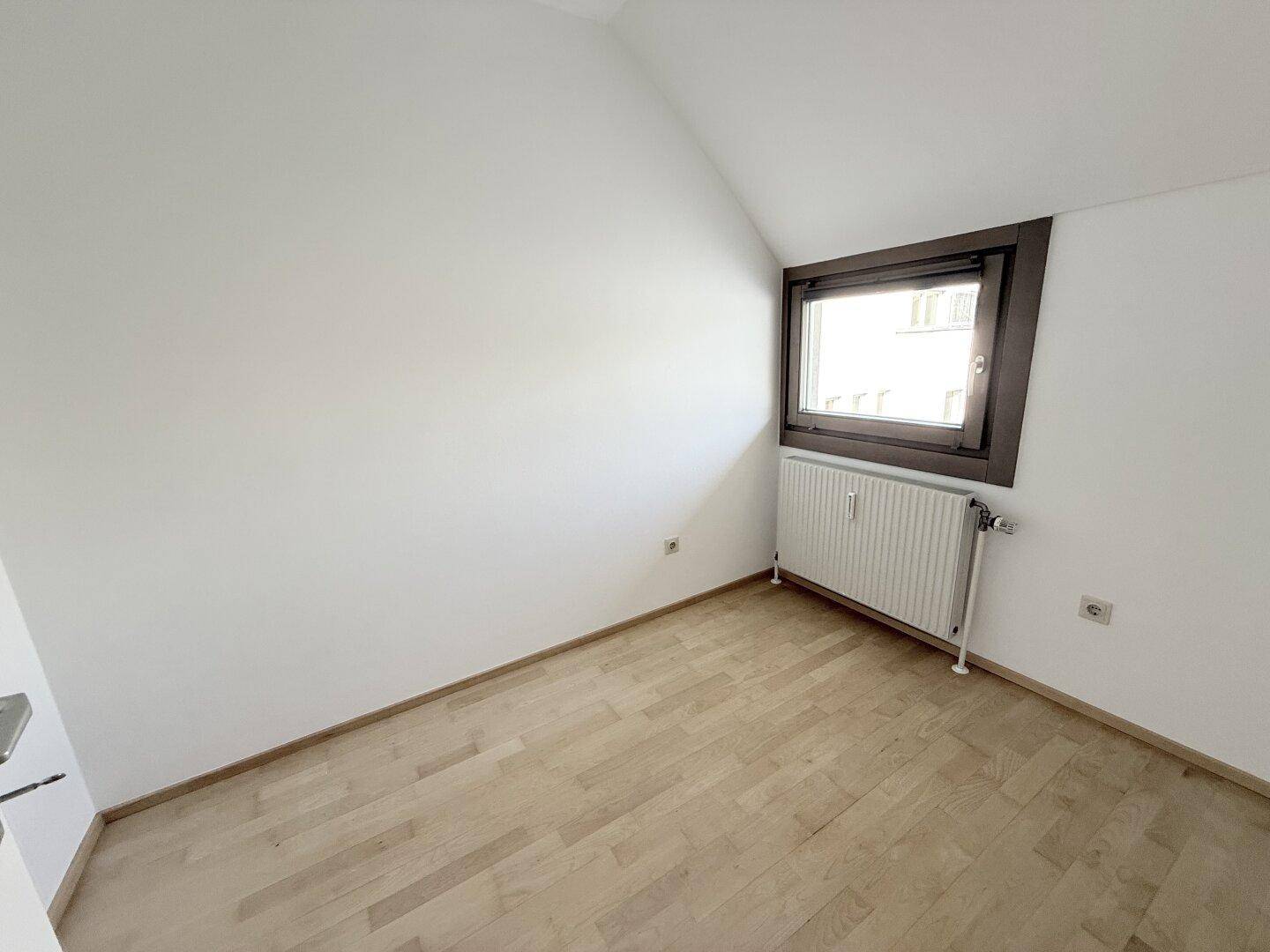 Gemütliches Zimmer mit hellem Holzparkett, Dachschräge und einem Fenster, das Tageslicht hereinlässt.