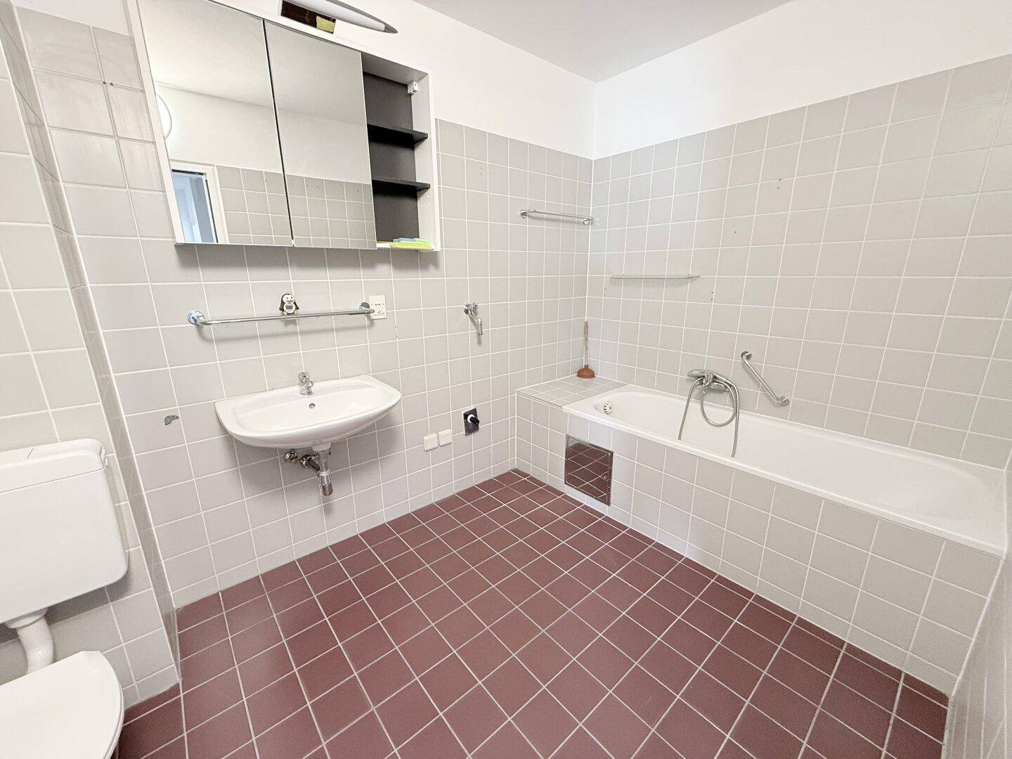 Badezimmer mit hellgrauen Wandfliesen, roten Bodenfliesen, Badewanne und Waschbecken mit Spiegel.