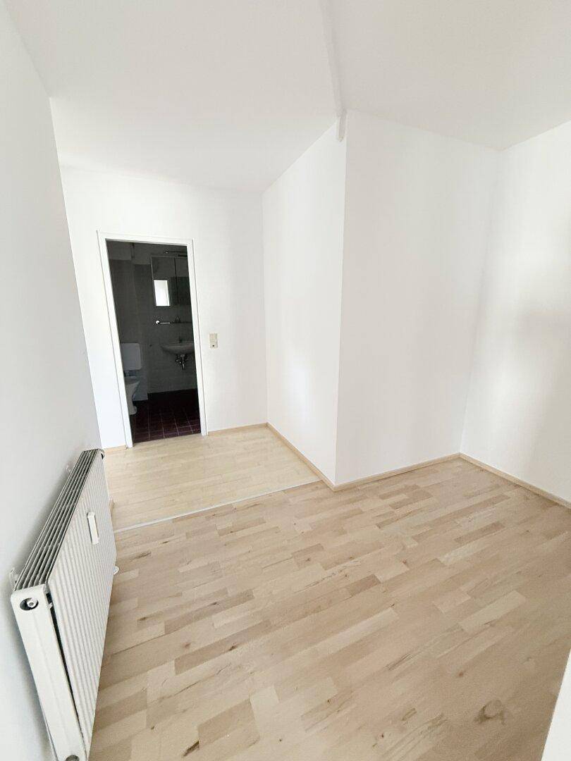 Helles Zimmer mit hellem Holzparkett und weiß gestrichenen Wänden, mit Zugang zum Badezimmer.