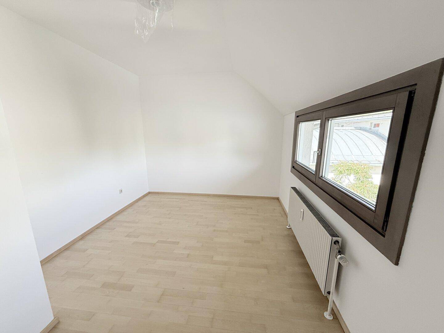 Helles Zimmer mit hellem Holzparkett, Dachschräge und einem Fenster mit Blick auf die Umgebung.
