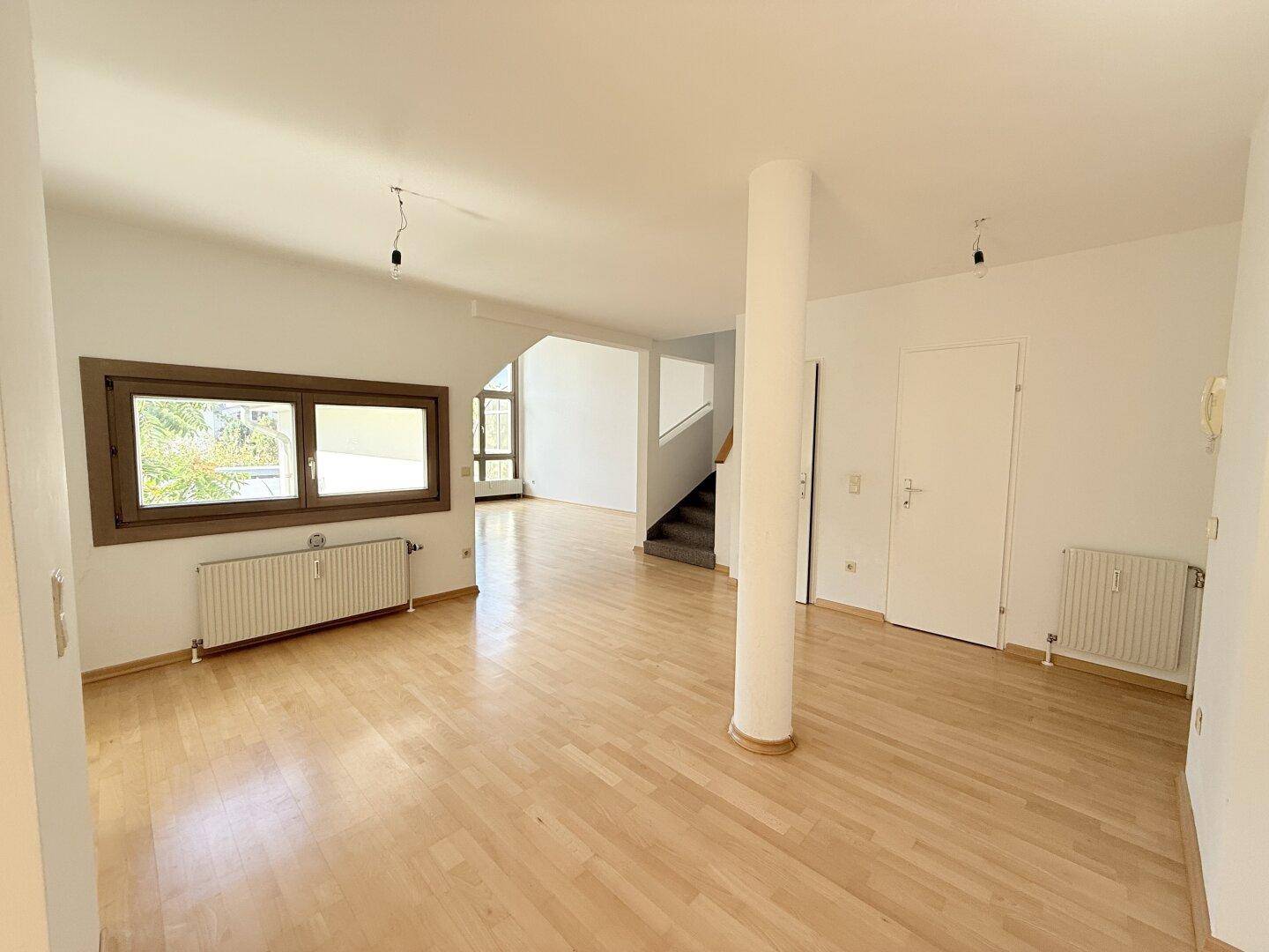 Geräumiges Wohnzimmer mit hellem Parkettboden, weiß gestrichenen Wänden und Blick auf den Treppenaufgang.