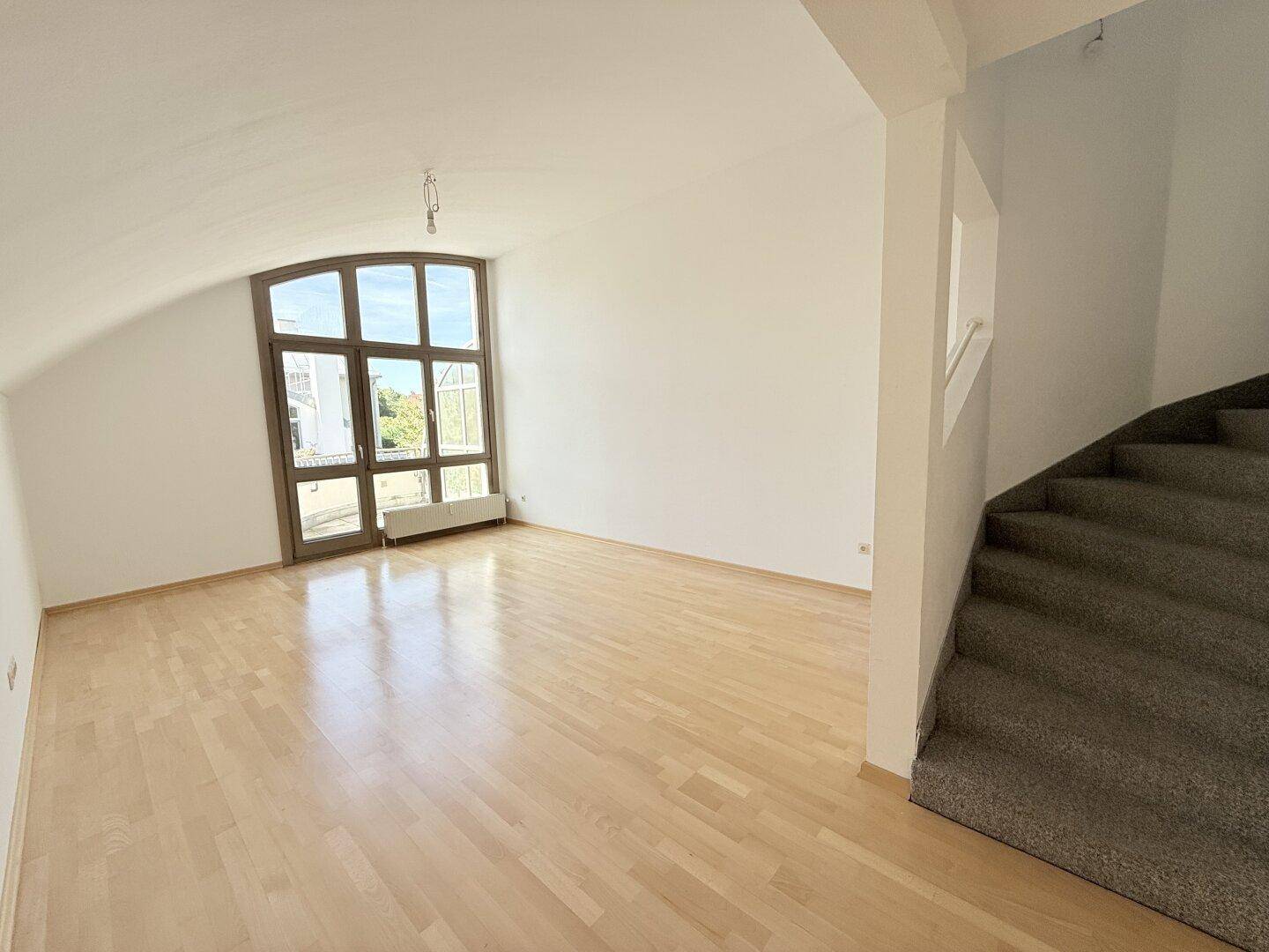 Heller Wohnbereich mit Parkettboden, großer Fensterfront und Zugang zu einer Treppe.