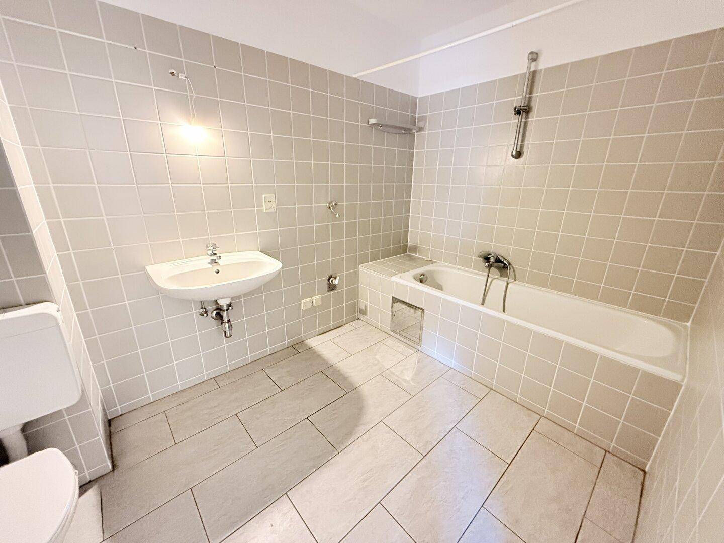 Badezimmer mit Badewanne, Waschbecken und hellgrauen Fliesen an Wänden und Boden.