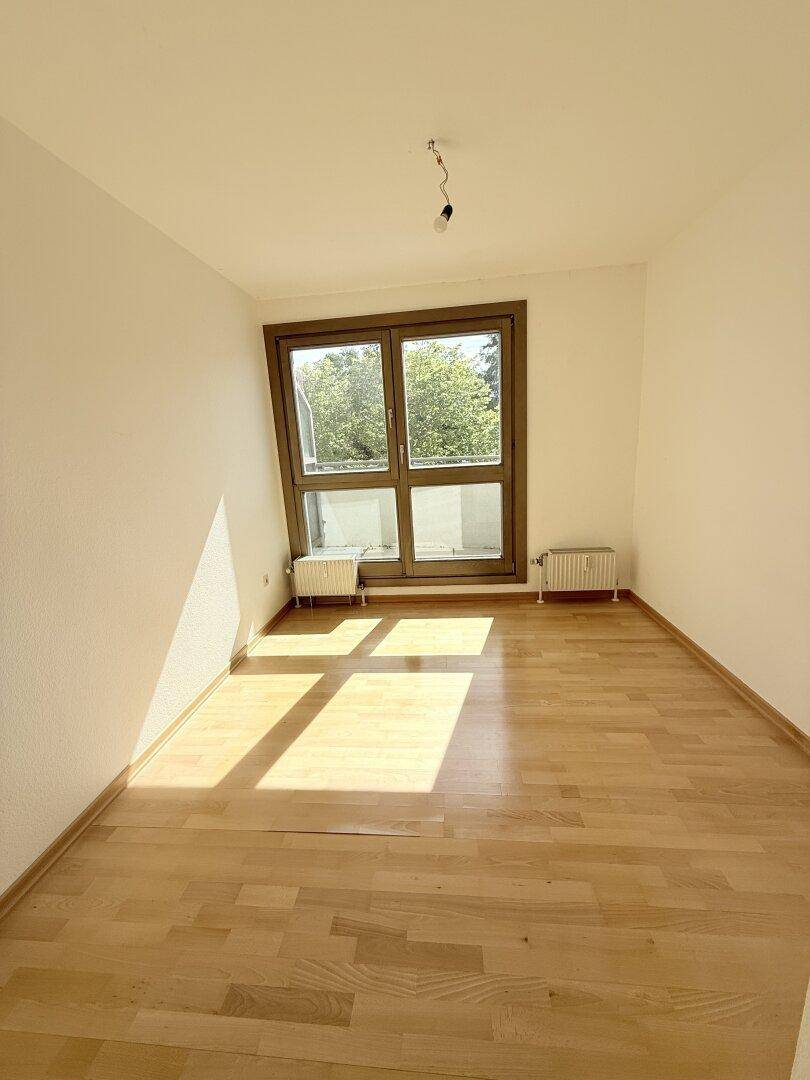 Helles Schlafzimmer mit Parkettboden und großem Fenster, das viel Sonnenlicht hereinlässt.