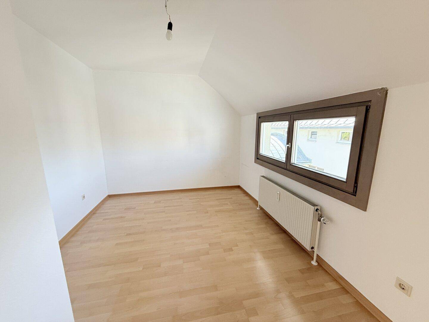 Schlafzimmer mit Parkettboden, schräger Decke und Fenster mit Blick nach draußen.